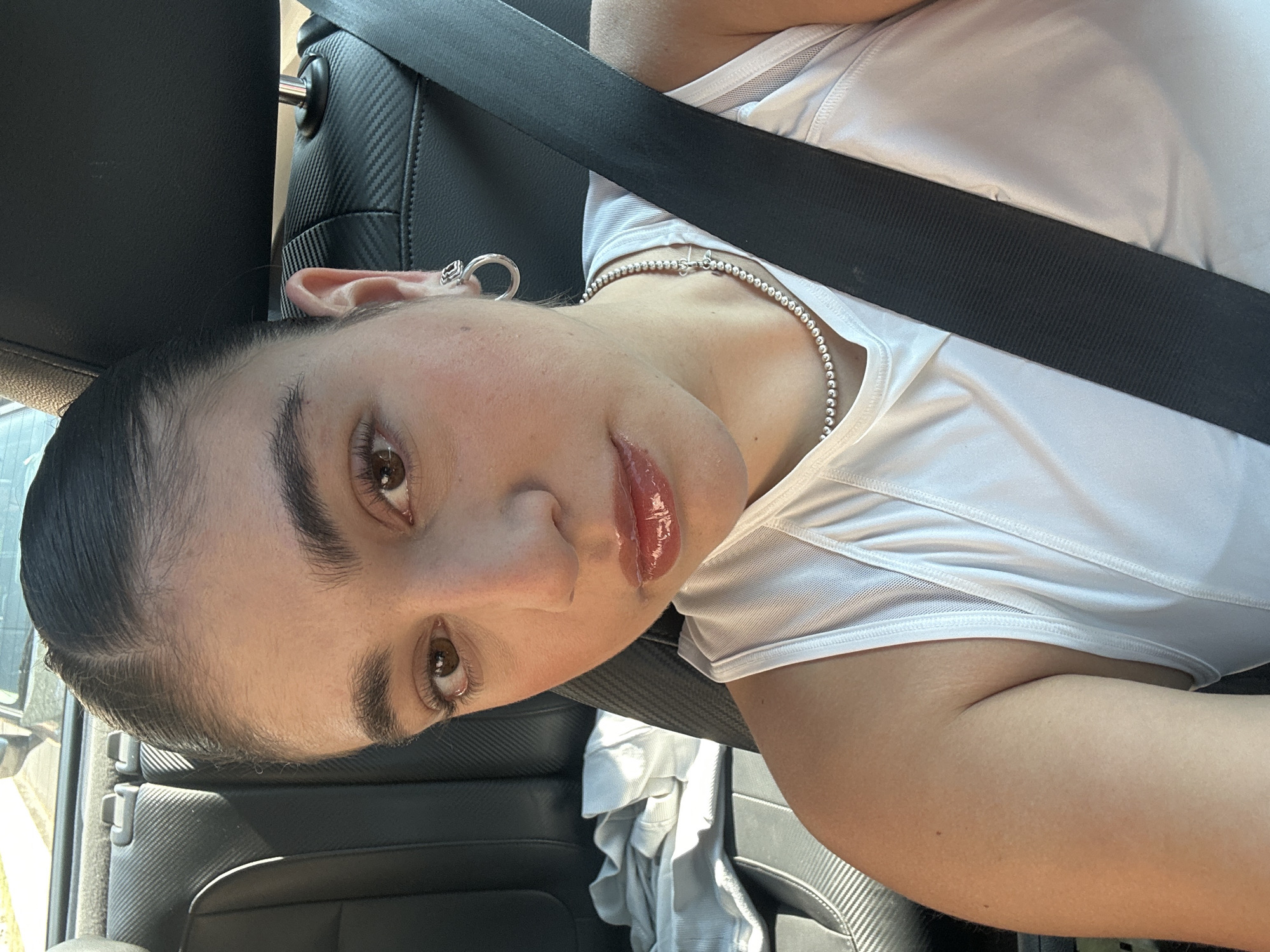 The image shows a person in a car wearing a seatbelt, a white shirt, and a silver ball chain necklace. The person has brown eyes, dark hair pulled back, and is wearing lip gloss. An earring is visible in the left ear. The background includes the car's interior, with a patterned headrest and door panel. No location information about San Bernardino Tlaxcalancingo, Mexico is discernible from this image.