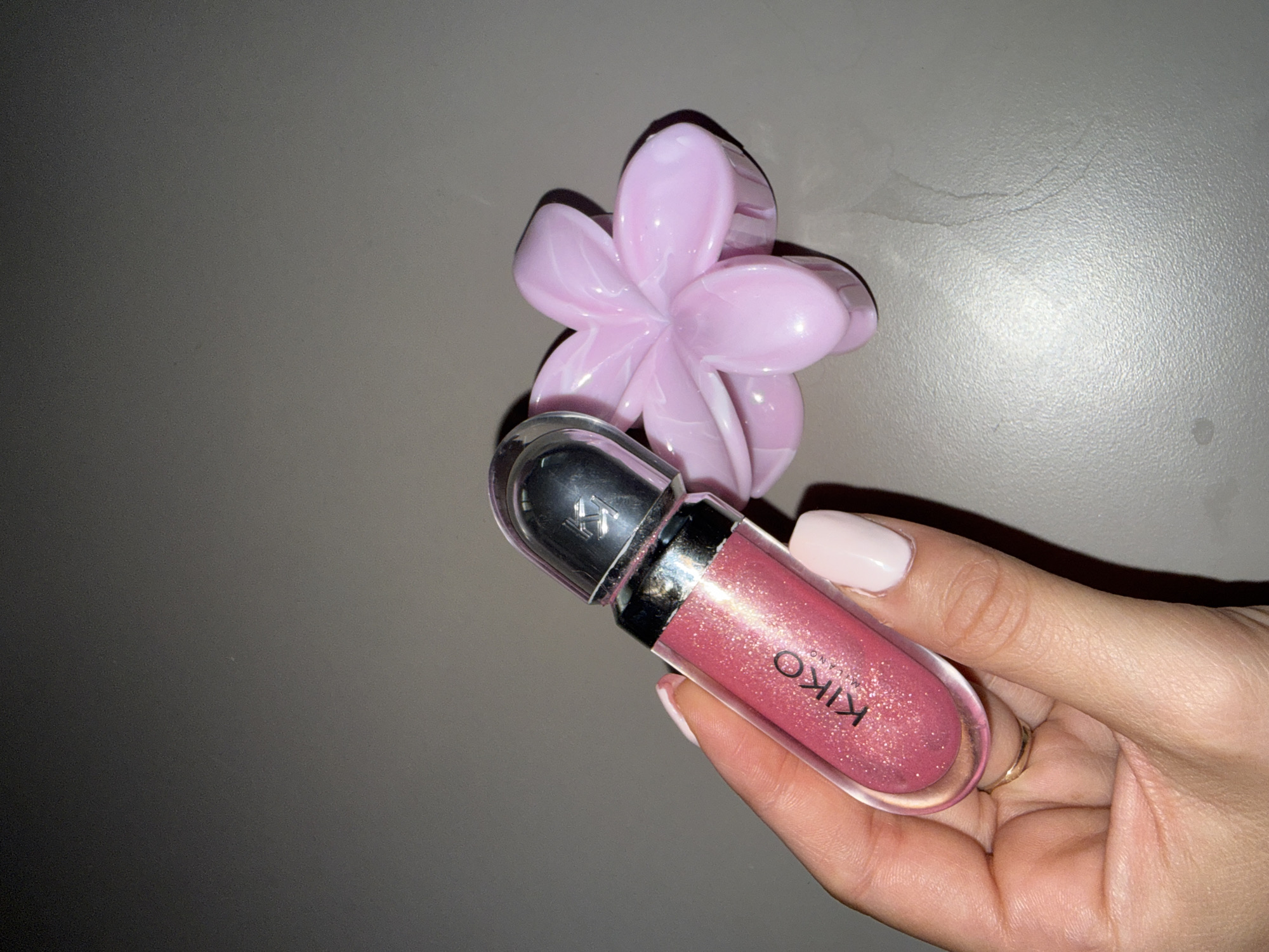 A close-up, eye-level shot shows a person's left hand holding a KIKO Milano lip gloss. The lip gloss tube is clear with a silver cap and contains a shimmering pink liquid. A pink, flower-shaped hair clip sits to the right of the lip gloss. The background is a textured, gray surface. The image was taken indoors.