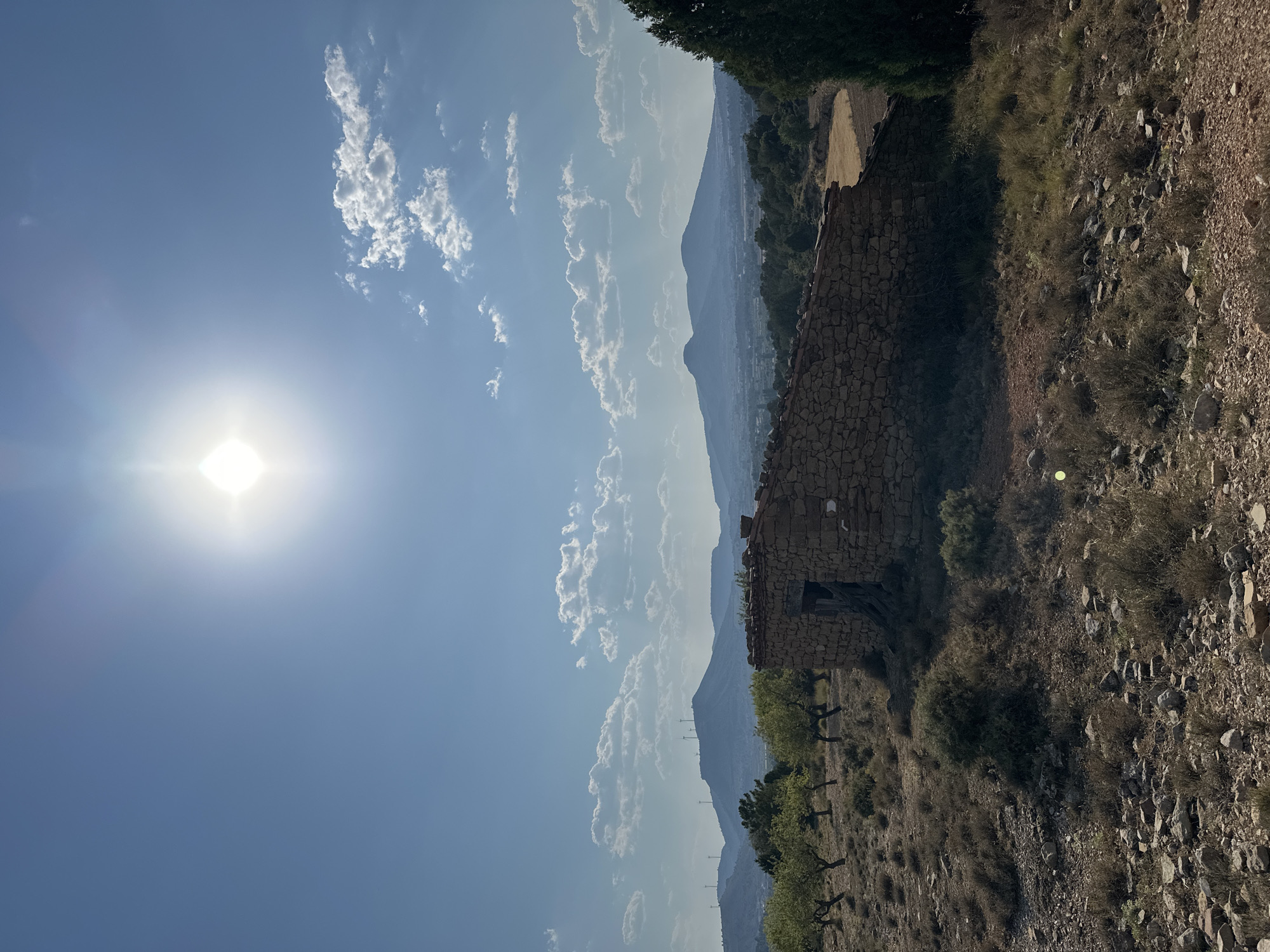 The image depicts a sun-drenched landscape with a clear blue sky and scattered white clouds. In the foreground, a dilapidated stone structure with an open doorway stands on a dry, rocky hillside dotted with sparse vegetation and a few scattered trees. In the mid-ground, a line of trees, possibly olive or almond trees, stretches across the frame, with a few more trees and dense brush visible on the right. In the background, rolling hills and mountains are silhouetted against the sky, with wind turbines visible on the distant peaks. A town or village is faintly visible in the valley. The overall scene suggests a rural, arid environment in Alpuente, Spain, under bright sunlight.