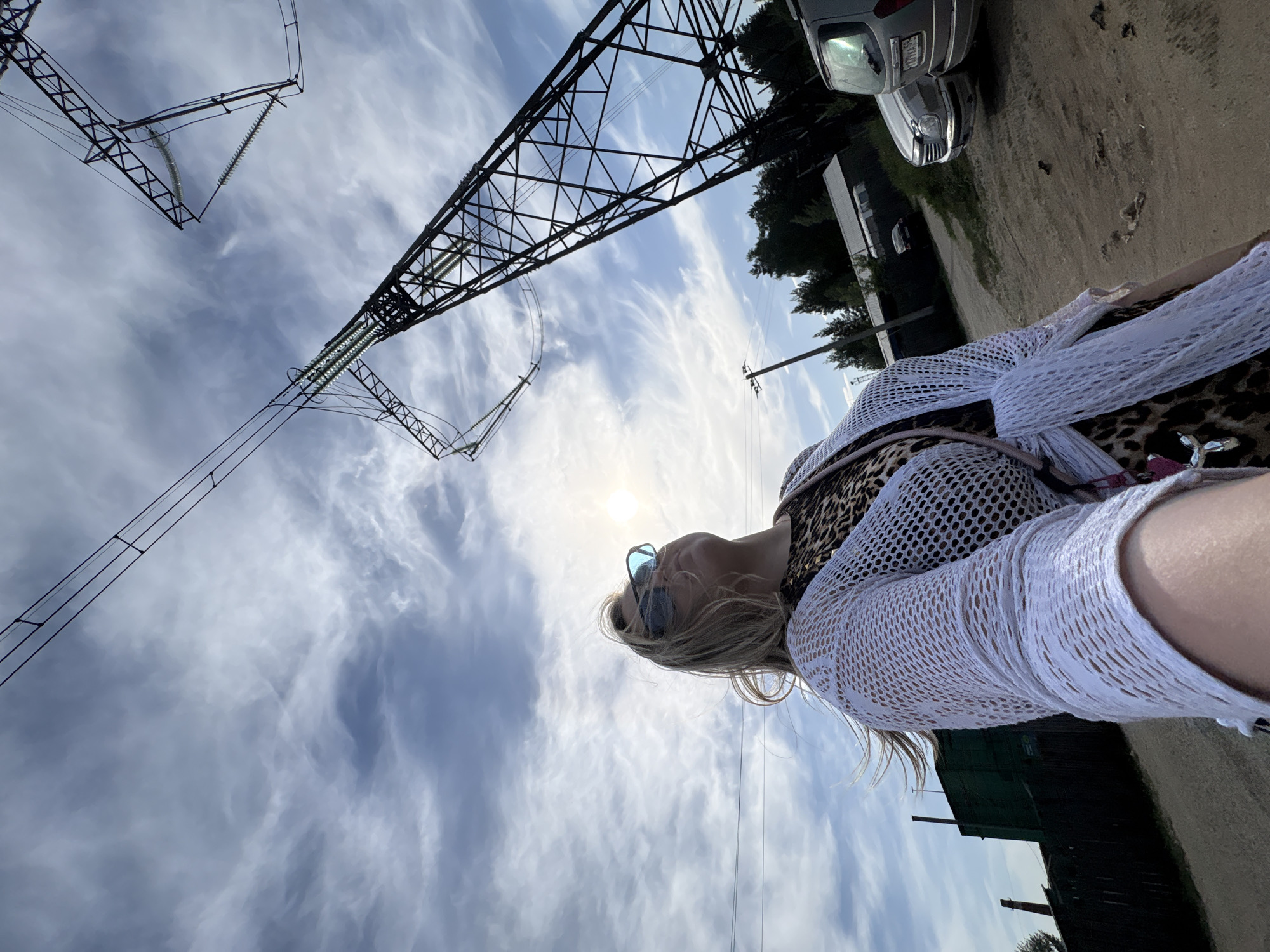 The image shows a woman with blonde hair and sunglasses, wearing a leopard print top and a white crocheted cardigan. She is standing in an outdoor area with utility poles and power lines overhead. In the background, there are trees, a vehicle, and buildings. The sky is partly cloudy. The location is near Noviy, Russia.
