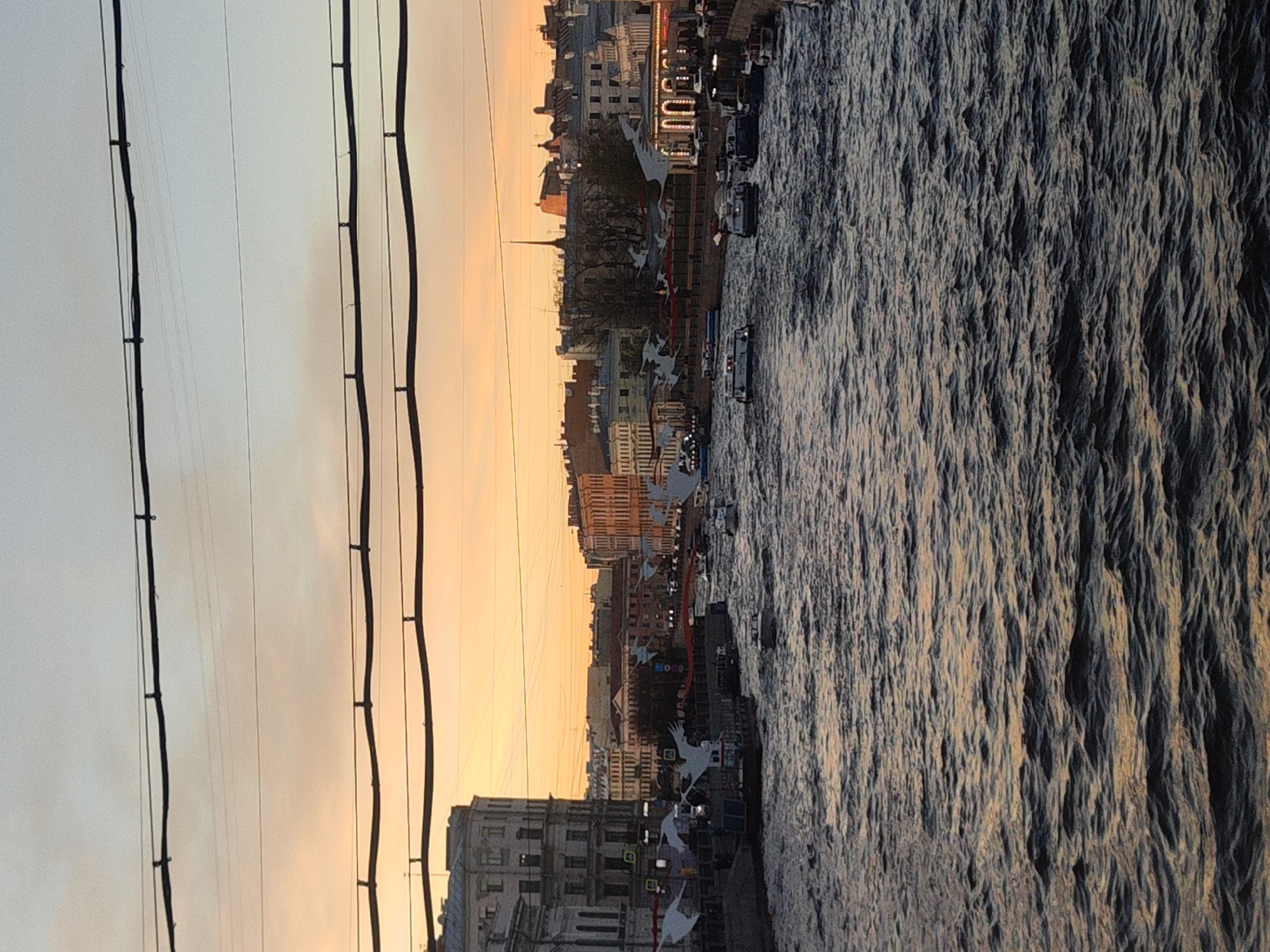 The image depicts a sunset over the Neva River in Saint Petersburg, Russia. The water is choppy with reflections of the setting sun. Along the embankment, buildings rise against the orange sky. There are a few small boats in the river. The presence of electric wires are visible in the upper left portion of the image. Decorative installations that include white birds are installed on the embankment.