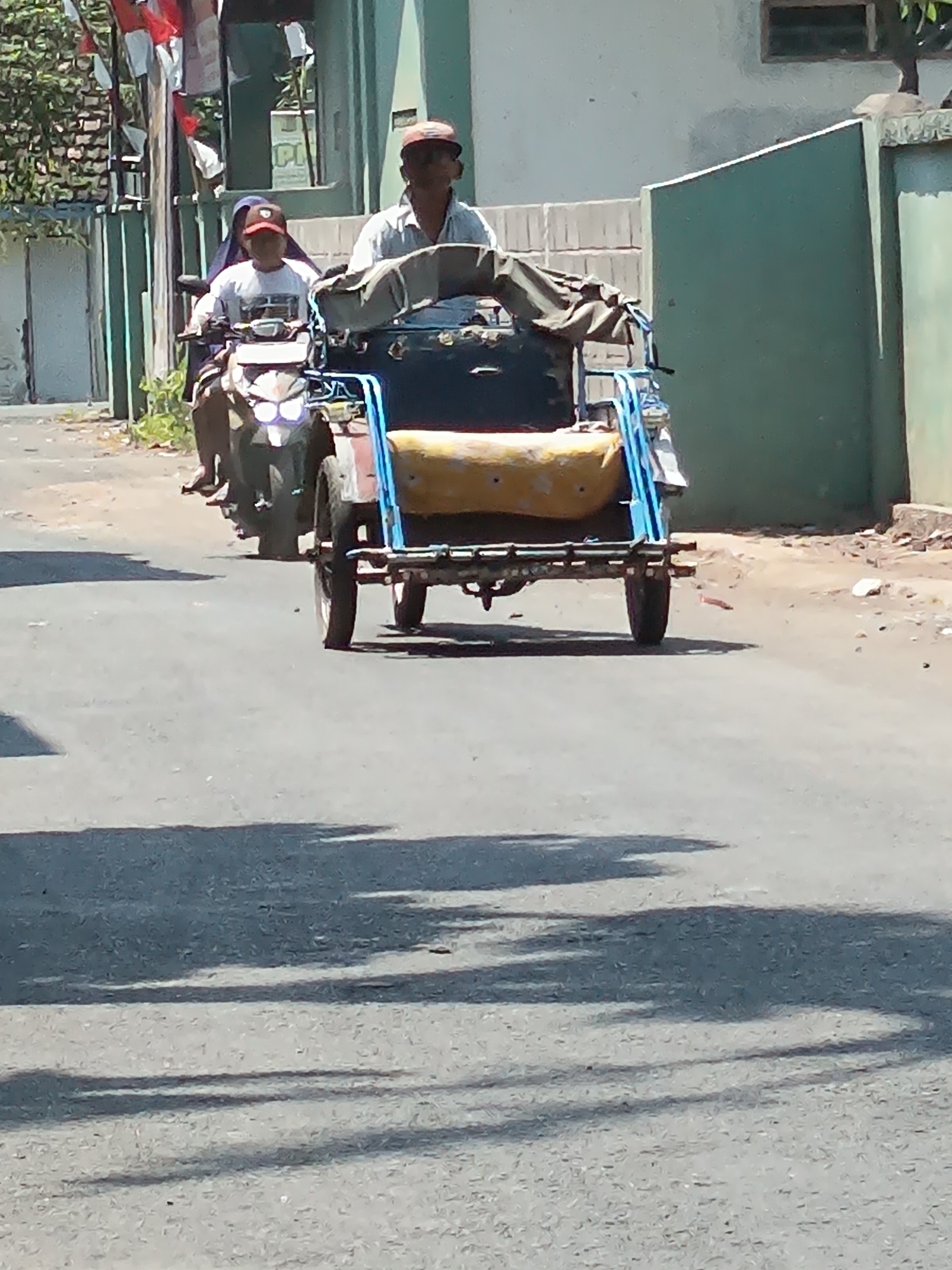 The image shows a street scene in Kandangjati Wetan, Indonesia, on a sunny day. Two people are visible on a motorcycle that is pulling a sidecar. The person in front is wearing a red cap and a light-colored shirt, and appears to be driving the motorcycle. Behind them, a person wearing a blue head covering and a red cap is seated in the motorcycle's passenger seat. The sidecar, which is blue with a yellow cushion, is empty. The motorcycle and sidecar are on a paved road. In the background, there are buildings and trees, and flags are visible hanging from a pole. The overall impression is of a typical street scene in a residential area.
