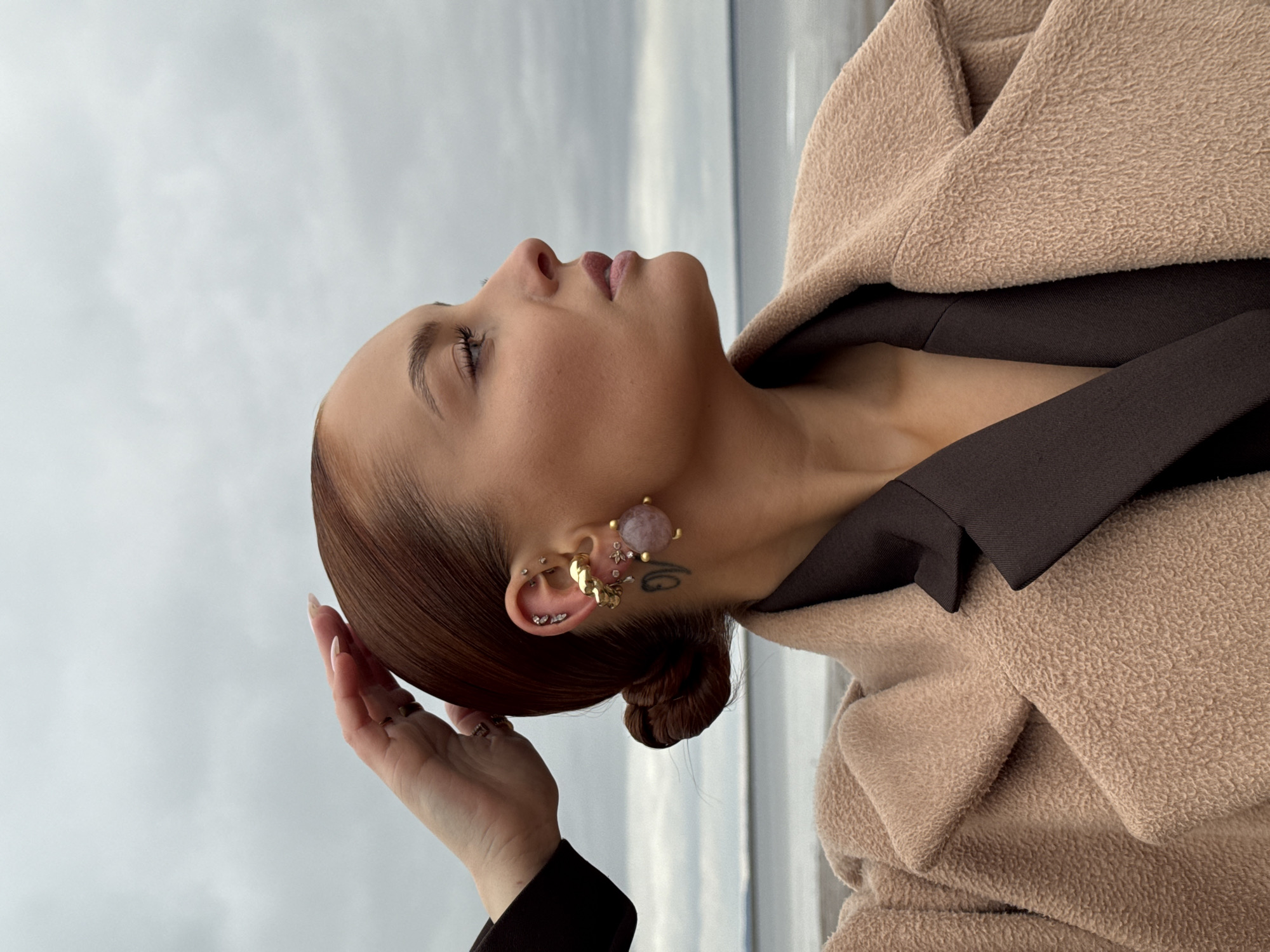A young woman is depicted in a side profile, looking upwards with her eyes gently closed or cast down, suggesting a moment of contemplation or peace. Her head is tilted back, revealing the contour of her neck. Her dark brown hair is styled in a low bun, and her left hand is lightly placed near the back of her head, touching her hair.
She wears a textured light beige or tan coat, appearing cozy and warm, layered over a dark brown top with a visible collar. Her left ear is notably adorned with multiple piercings: a large, striking earring featuring a pinkish-purple stone set in gold, along with a gold ear cuff and several smaller gold and silver studs. A small, dark tattoo is visible on her neck, just behind her left ear.
The background shows an overcast sky, with a faint horizon line hinting at a body of water or distant landscape. The lighting is soft and diffused, characteristic of a cloudy daytime setting. The overall atmosphere is tranquil. There is no visible text. The location context for this scene is Chaikovsky, Russia.
