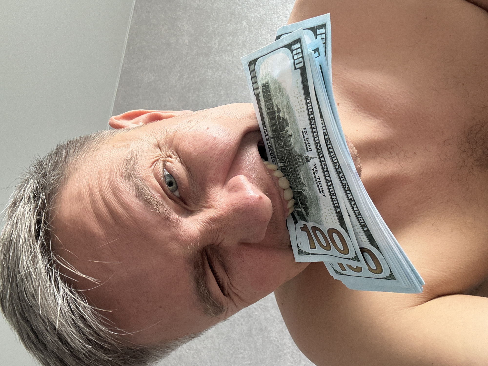 A photograph shows a person with gray hair, holding a stack of U.S. one hundred dollar bills in their mouth. The person's torso is visible, suggesting a shirtless appearance. The background appears to be a plain, light-colored wall. The image's orientation is rotated, giving a tilted perspective of the subject. The photograph was taken in Бараўлянскі сельскі Савет, Belarus.