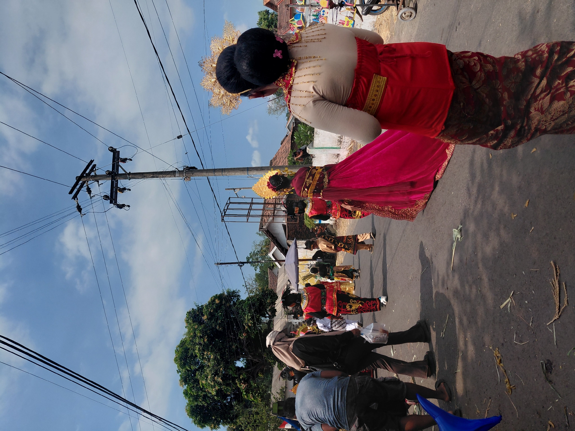 In Bimo, Indonesia, a procession takes place on a paved street under a blue sky with scattered clouds. Overhead, a network of black electrical wires crisscrosses the sky, supported by a wooden utility pole. A large, leafy green tree stands to the left of the frame.
Several individuals are participating in the procession. In the foreground, a person is viewed from behind, wearing a red garment over a patterned skirt and an ornate golden headdress. To their left, another individual in a vibrant pink gown with gold embellishments and a golden headdress walks away from the camera. Behind them, a line of participants in traditional red and gold attire, some with head coverings, proceed down the street. Among them are individuals in more casual clothing, and a person carrying a plastic bag. The street is lined with houses and a white wall, with some decorative items visible on the right side. Debris, including dried leaves and straw-like material, is scattered on the asphalt.