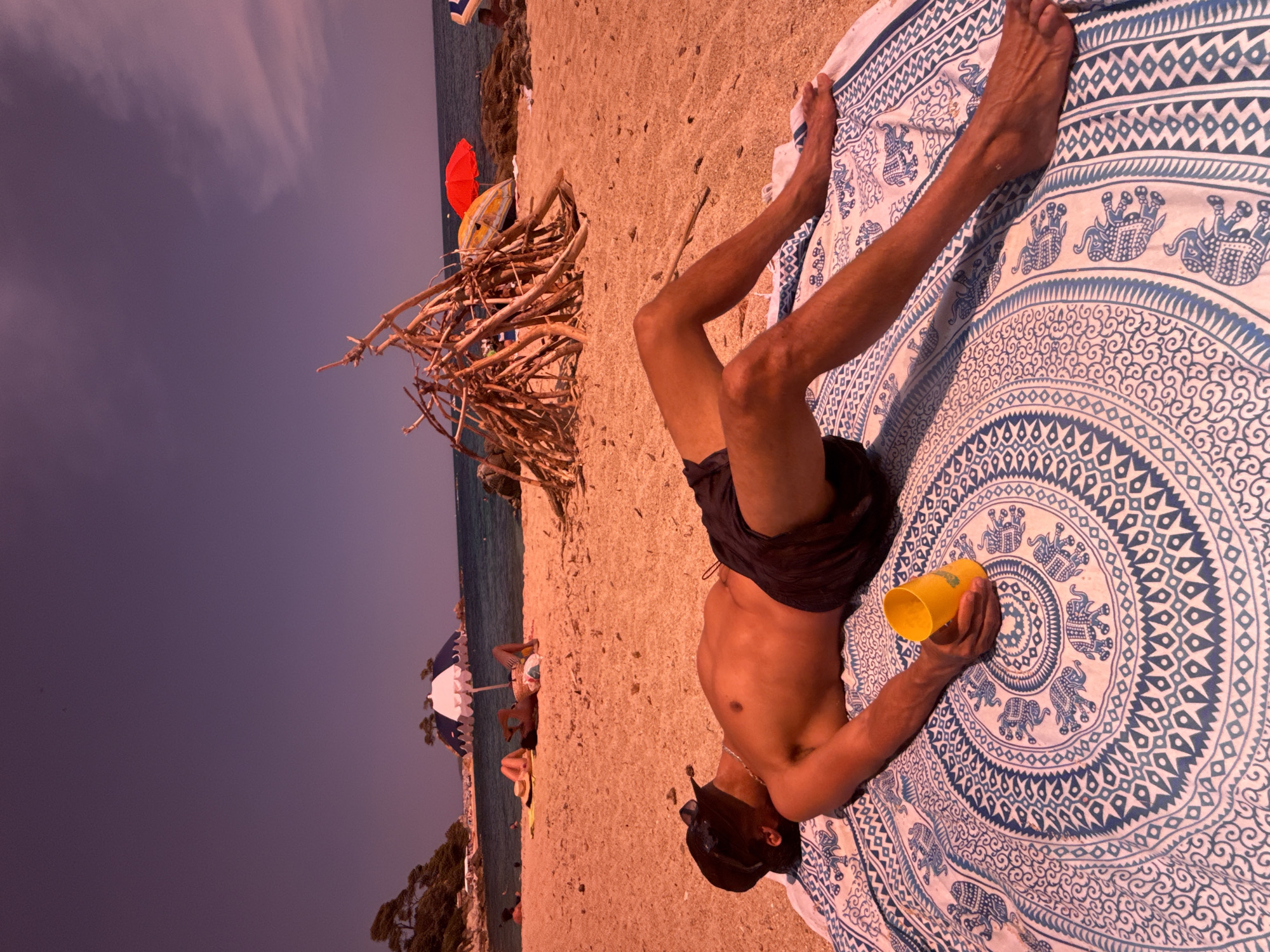 In Calonge i Sant Antoni, Spain, on a sunny beach, a man lies on his back on a circular, blue and white patterned towel. He wears a black baseball cap and dark swim trunks. A yellow cup rests in his hand. In the background, other beachgoers are visible, some under umbrellas, and a structure made of driftwood stands on the sand. The sea meets the horizon under a purple-tinted sky.