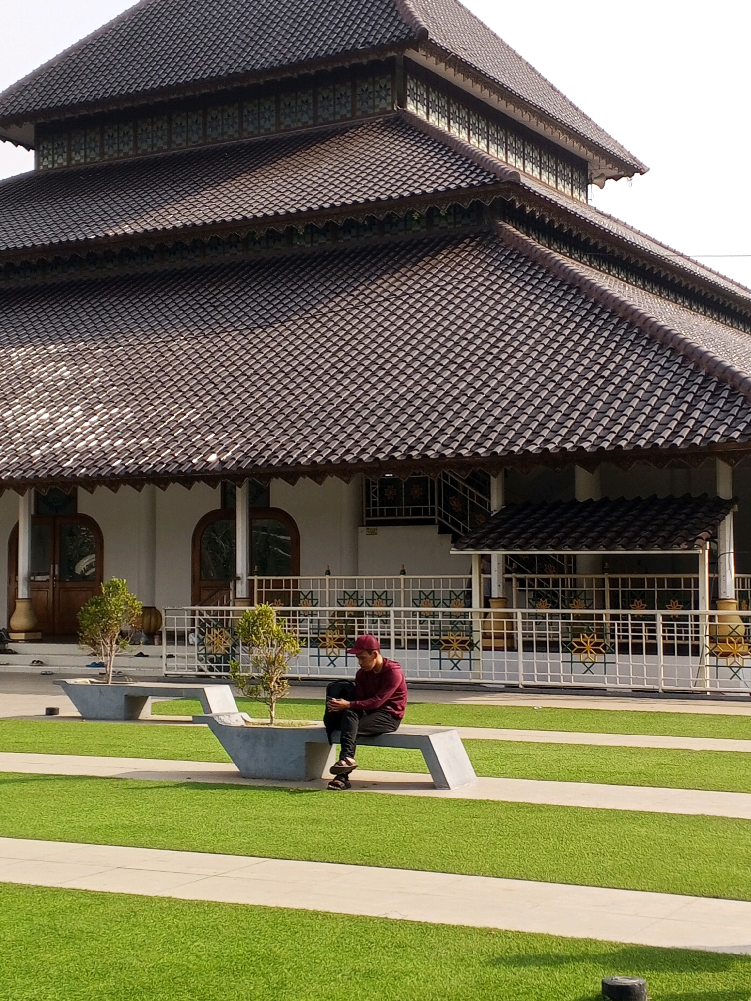 In the city of Serang, Indonesia, a young man wearing a maroon long-sleeved shirt and a maroon cap sits on a concrete bench in a park-like area in front of a mosque. He is looking down at a mobile device in his hands. The mosque features a distinctive multi-tiered roof with dark brown tiles and intricate decorative elements along the eaves. The building has white walls and arched wooden doorways. A white metal fence with decorative patterns surrounds the mosque's grounds. The park area is characterized by neatly manicured green grass and paved pathways. Two small, potted trees are planted in concrete planters on either side of the bench. The scene is bathed in sunlight, casting shadows on the grass.
