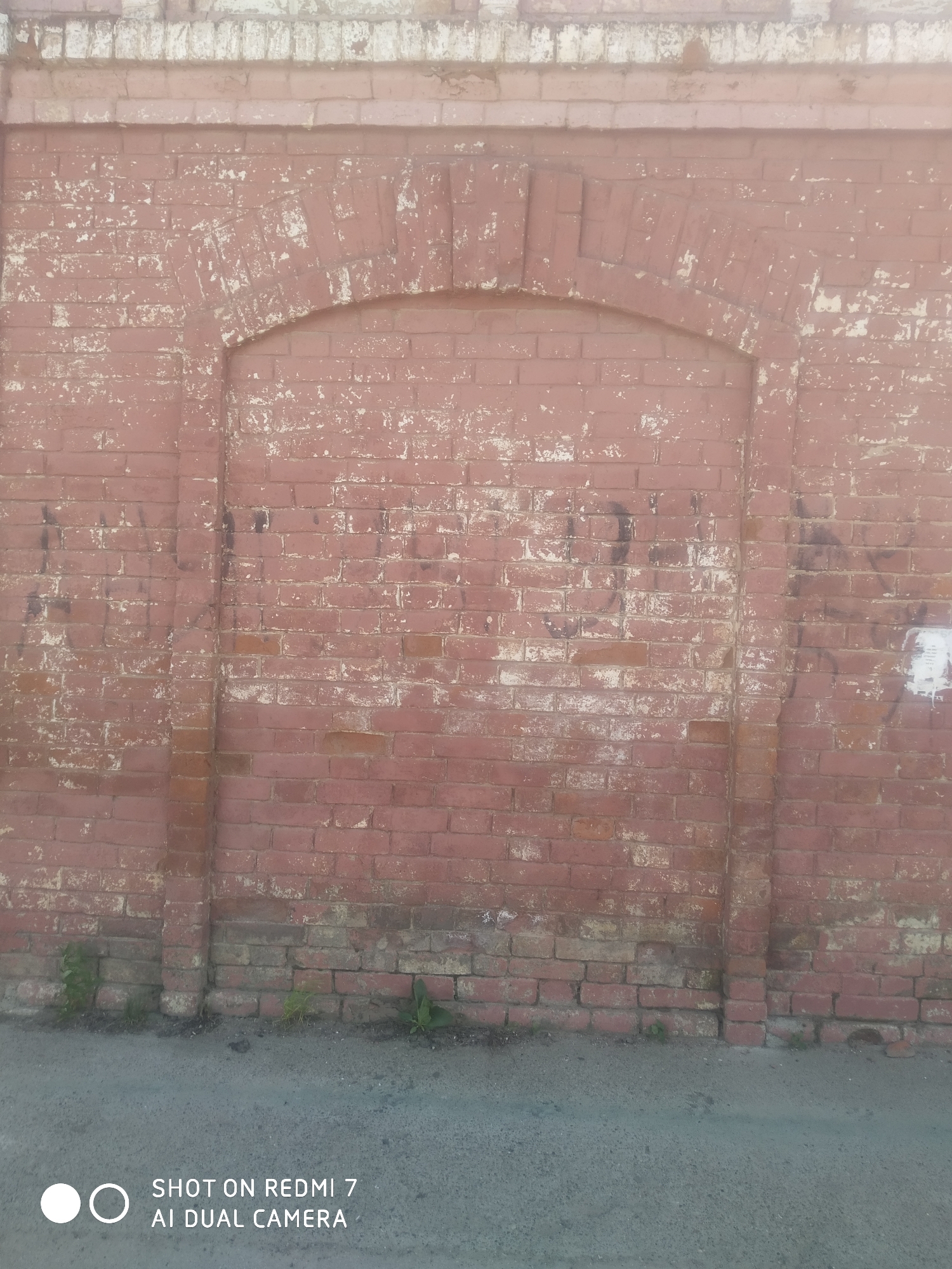 A brick wall with a filled-in arched doorway in Ochansk, Russia. The wall is made of reddish-brown bricks with patches of white discoloration. The arched doorway is bricked in and has a rounded top. The base of the wall is partially obscured by weeds. A gray asphalt surface is visible at the bottom of the image.