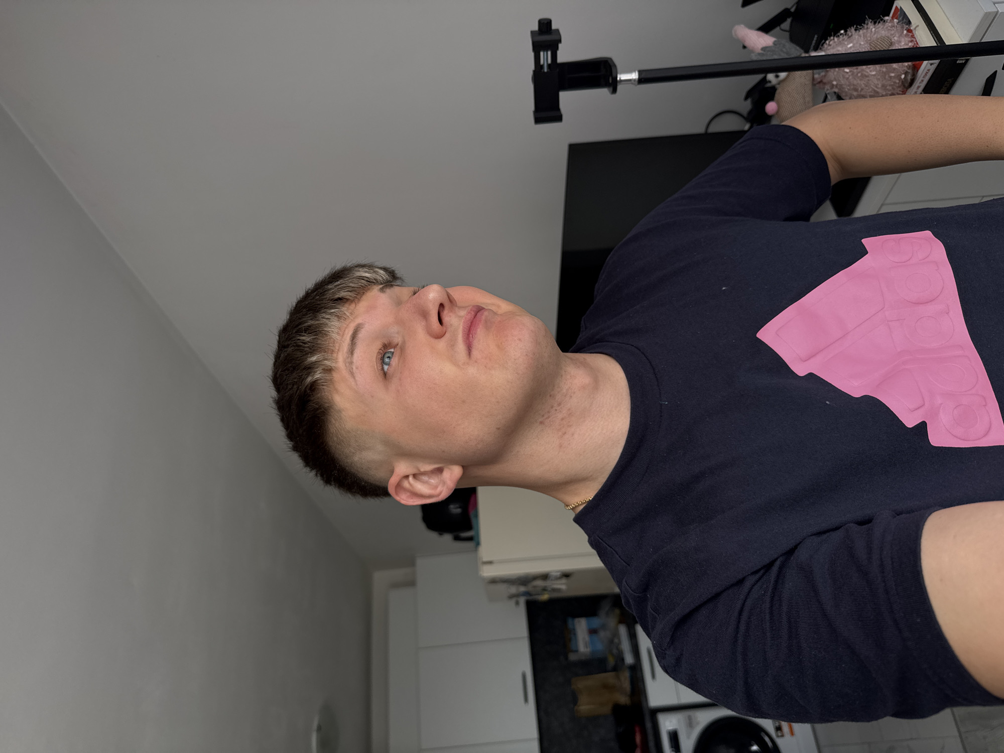 A fair-skinned male with short, blond-tinted hair and blue eyes is angled upwards, facing right. He wears a dark-colored shirt with a large, light pink logo. Part of a microphone stand is visible above him, as well as part of a desk, books, and a plush animal. Behind him and to the left is a wall and kitchen with a washing machine. The location in the city of Plovdiv, Bulgaria cannot be determined from the image.