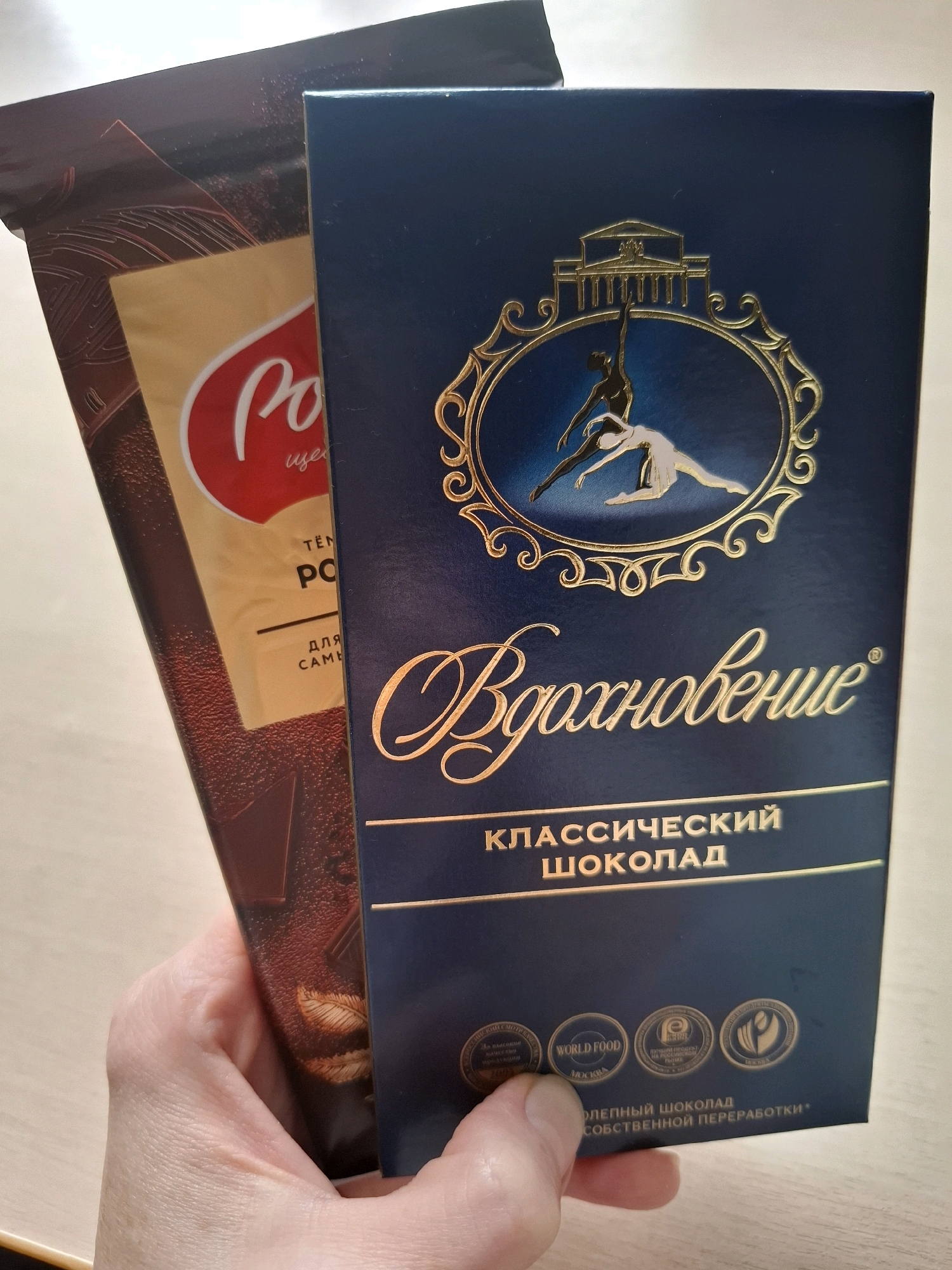 Two chocolate bars are held in a person's left hand. The bar in the foreground is dark blue with gold lettering and an emblem of two dancers within an ornate frame. The text on this bar reads "Вдохновение" (Inspiration) and "КЛАССИЧЕСКИЙ ШОКОЛАД" (Classic Chocolate). Below this are four circular seals. The bar in the background is brown and gold with red accents. It appears to be a dark chocolate bar with imagery of chocolate pieces and leaves. The provided image does not contain any information that allows for the determination of its location in the city of Tyumen, Russia.