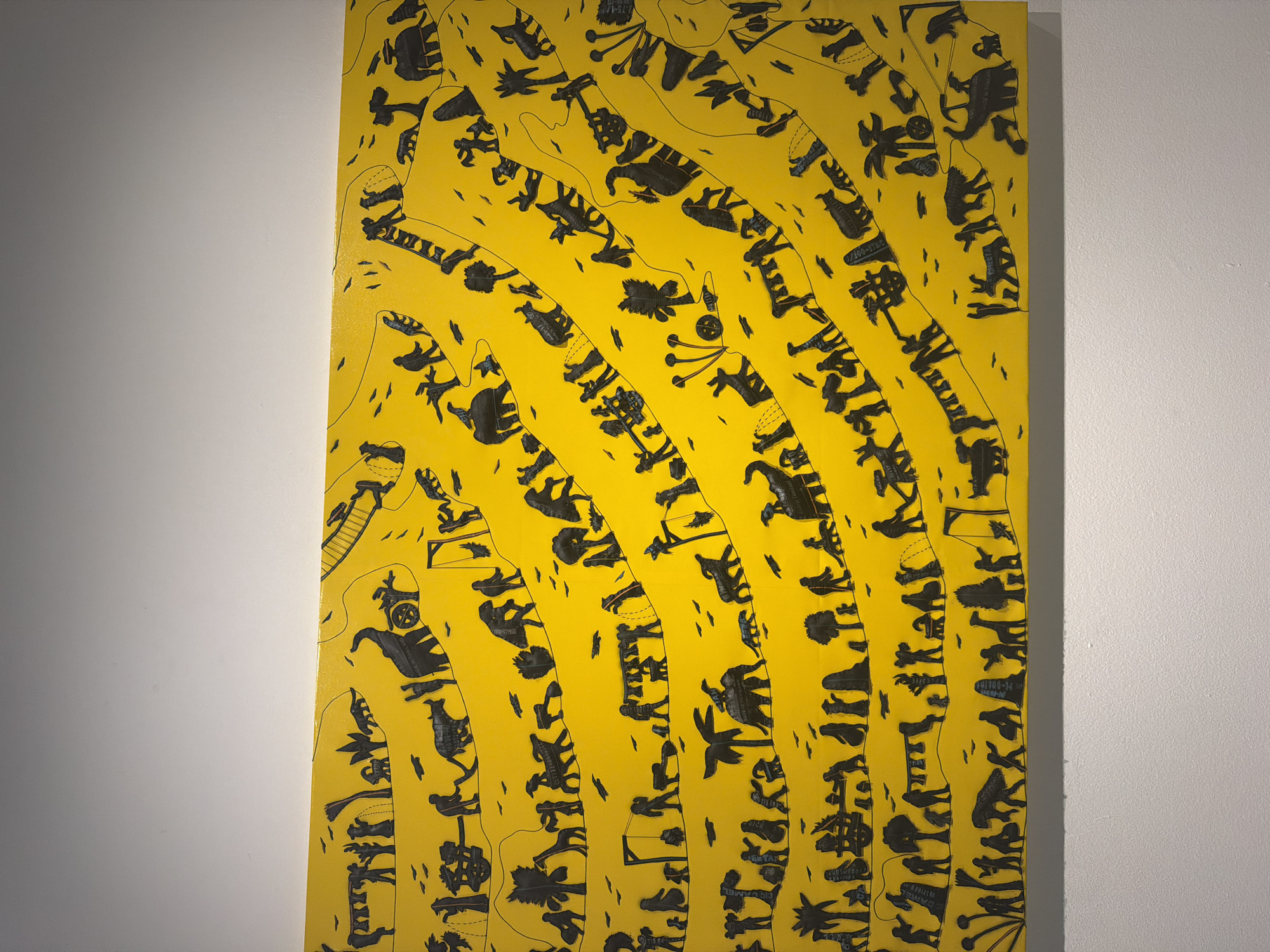 The image is a close-up, eye-level shot of a yellow canvas painting with black silhouettes of figures and animals. The painting depicts a winding, layered scene filled with various figures engaged in different activities and accompanied by animals, set against a vibrant yellow background.
In the foreground, there are rows of silhouettes arranged in curved bands. These figures include humans, some carrying objects or pulling carts, and various animals such as elephants, giraffes, cows, and what appear to be wolves or dogs. Some human figures are shown interacting with the animals, while others are engaged in solitary tasks like hanging laundry or tending to plants. The environment is suggested through stylized trees, some with palm fronds, and abstract plant-like forms.
The arrangement of the silhouettes creates a sense of movement and narrative across the canvas, with some elements appearing to move uphill or downhill. There are also subtle details like small birds in flight and abstract shapes that might represent ground or texture. The overall impression is one of a bustling, albeit stylized, depiction of life and nature.
This artwork appears to be displayed on a white wall, with the bottom edge of the painting just above the base of the wall. The lighting suggests an indoor setting, possibly an art gallery or exhibition space. There is no indication within the image itself of its specific location in Bangkok, Thailand, or any other city.