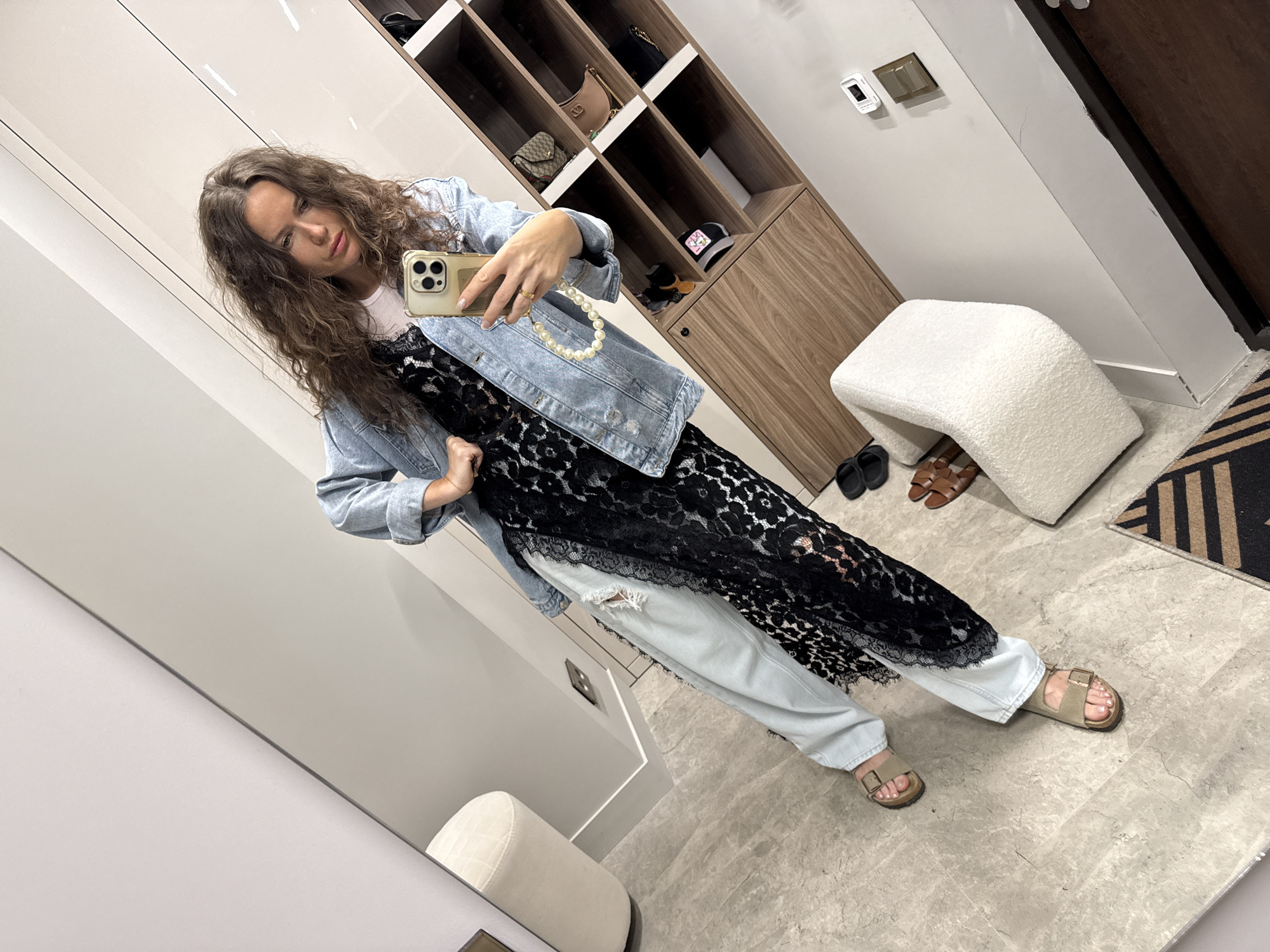 A woman takes a selfie in a room with a mirror. She is wearing a light blue denim jacket over a black lace dress and light blue ripped jeans. She has a pearl phone strap attached to her phone. Her feet are clad in tan slide sandals. The background features a large mirror reflecting the scene, a closet with shelves displaying handbags, and a white boucle armchair.