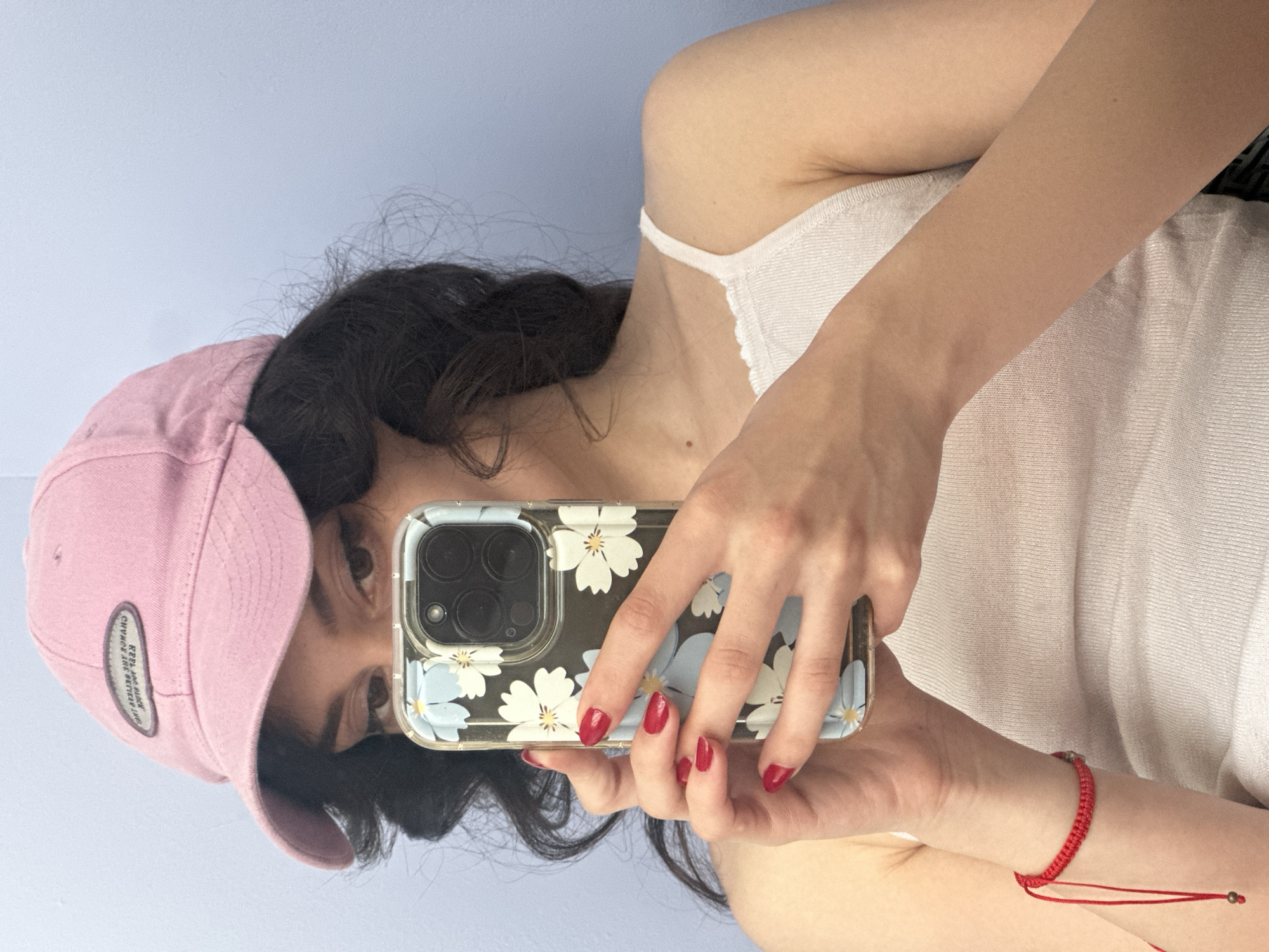 A young person with dark, wavy hair, wearing a pink baseball cap and a white tank top, holds up a smartphone with a floral case to take a selfie. Their nails are painted a bright red. On their left wrist, they wear a thin red string bracelet with a small, dark bead. The background is a plain, light blue wall. The image appears to be taken indoors, likely in a residential setting. There is no indication of the location being in the city of Sofia, Bulgaria.