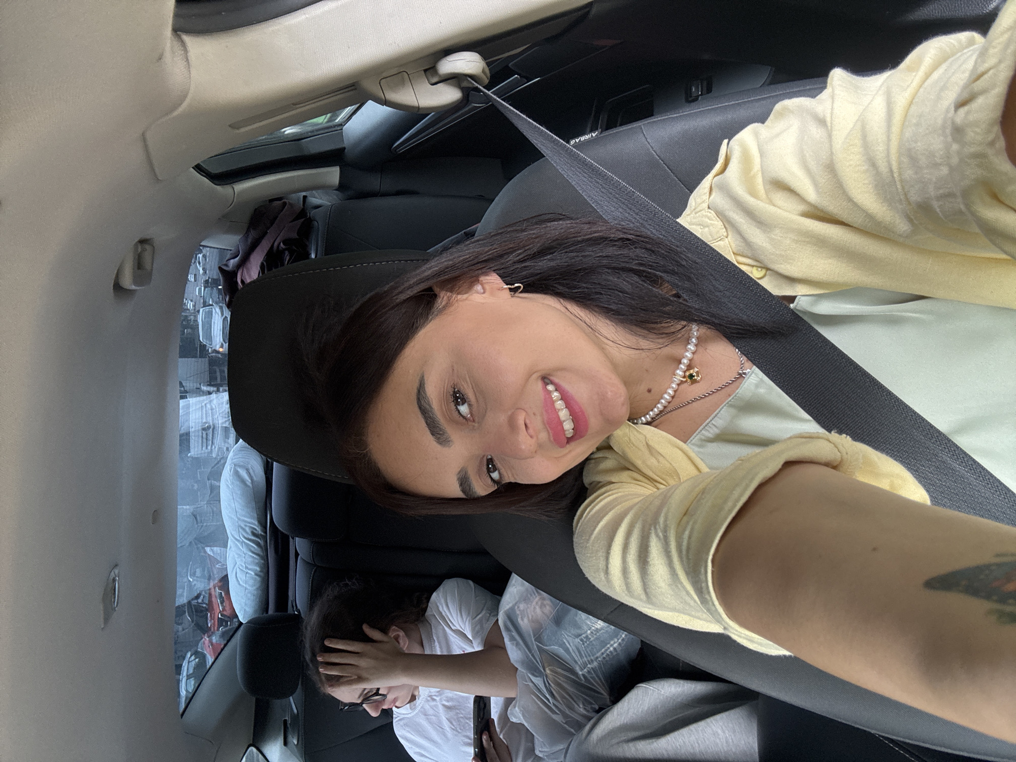 In the image, two individuals are seated inside a car. The person in the foreground, on the right, is a woman with dark hair, wearing a light yellow long-sleeved shirt and a pearl necklace. She is smiling at the camera, and her right arm, adorned with a tattoo, is visible. The other individual, a woman with dark hair and glasses, is in the back seat on the left, partially obscured by her hand covering her face. She is wearing a white t-shirt and jeans.
The scene appears to be daytime, with light filtering in from the car's sunroof, indicating it is likely not nighttime. The interior of the car, with black leather seats and a grey headliner, is visible. Outside the car windows, a street scene with other vehicles is discernible, suggesting an urban environment, possibly Tbilisi, Georgia, as indicated by the location context. The overall mood seems relaxed and casual, with the smiling subject conveying a positive emotion.