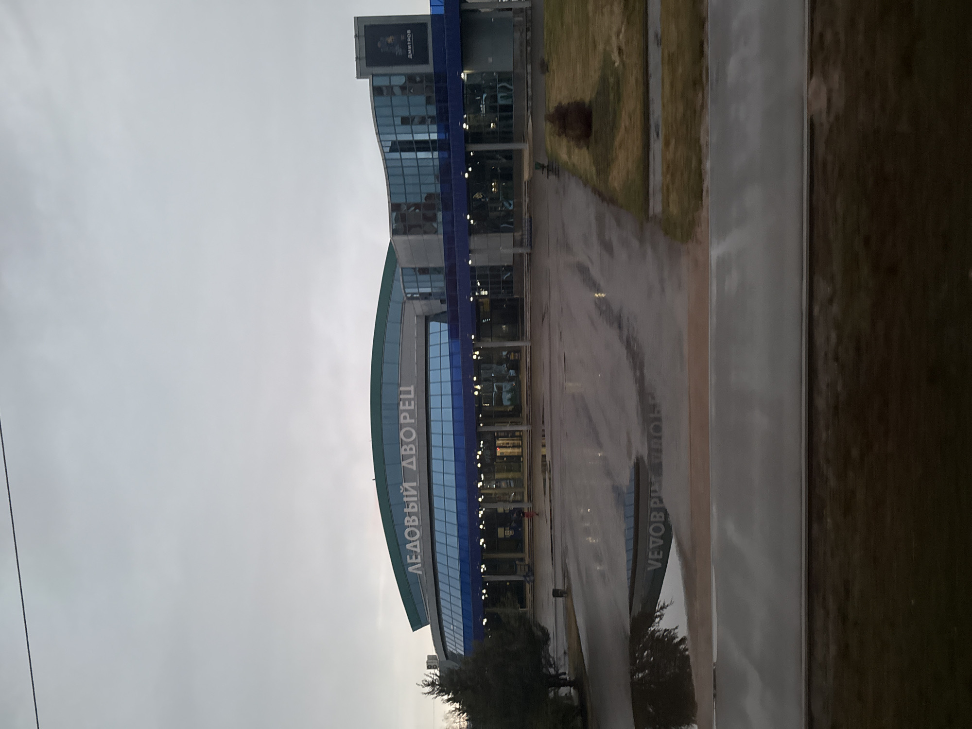 The image shows the Ice Palace "Dmitrov" in the city of Dmitrov, Russia. The building is a modern structure with blue and glass facade. The sky is overcast and the ground appears wet, suggesting recent rain. Trees are seen near the building. The surrounding area appears to be a paved area with grass.