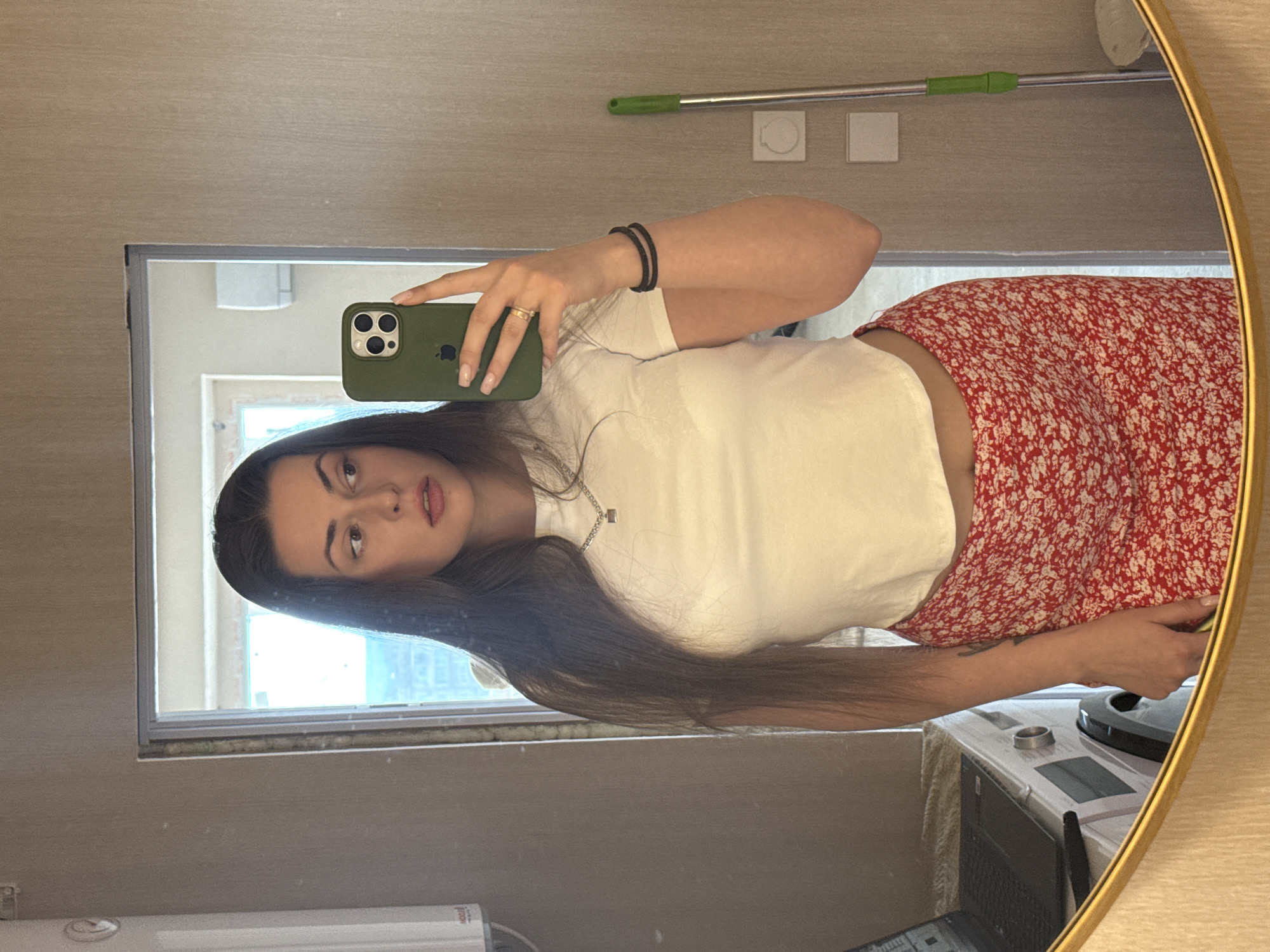 A woman with long, dark hair and fair skin takes a mirror selfie. She is wearing a white, cropped t-shirt and a red skirt with a white floral pattern. She has on a silver necklace with a small rectangular pendant, and a ring on her right ring finger. Her right arm is bent at the elbow, and her hand is holding a green smartphone. On her left wrist, she has two black rubber bracelets. The background shows a doorway, a washing machine with a laptop on top, and a mop leaning against the wall. The lighting is bright, likely from a window visible in the reflection.