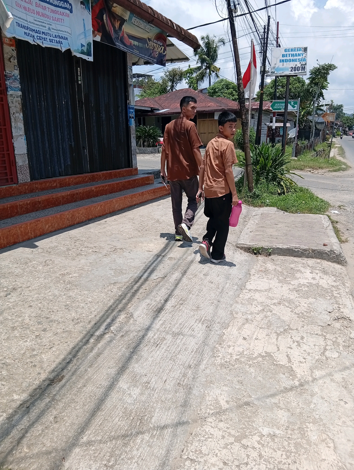 Two individuals, likely father and son, walk on a concrete sidewalk adjacent to a street. The older individual wears a brown t-shirt and dark pants, while the younger wears a light brown t-shirt and black pants, carrying a pink water bottle. They are walking away from the camera. In the background, a commercial building with closed metal shutters is visible, alongside a street with traffic and vegetation. A sign for "Gereja Bethany Indonesia" with an arrow indicating a distance of 200 meters is posted on a pole, suggesting the location is within the City of Gunungsitoli, Indonesia. An Indonesian flag and an evacuation route sign are also present. The scene is illuminated by daylight, casting shadows on the sidewalk.