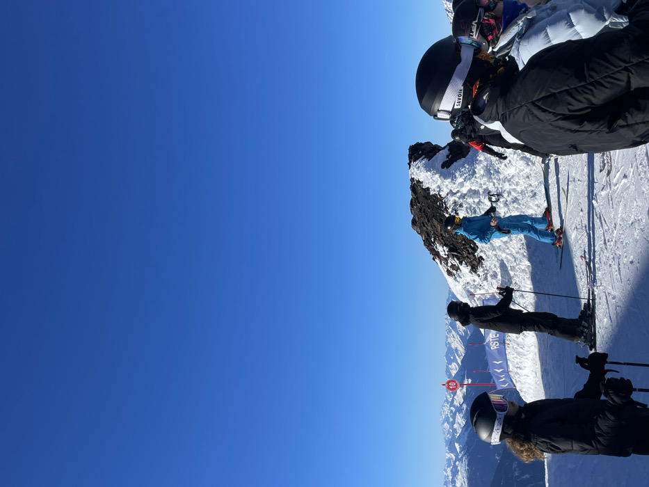 The image captures a vibrant, sunny day on a snow-covered mountain ski slope in Séez, France, showing at least five skiers paused amidst a scenic alpine landscape.
In the foreground, a skier in a black jacket and a black helmet with white goggles, featuring "SAVOIE" text on the strap, is visible from behind, holding ski poles. Another skier in a dark jacket and helmet is positioned just behind them. Further into the mid-ground, a skier in a striking bright blue ski suit and a yellow helmet stands on skis, situated next to a rugged, snow-covered rocky outcrop. Additional skiers are partially visible in the background.
The setting is a high-altitude ski resort, characterized by well-groomed snow, ski equipment, and resort infrastructure. A red marker post with the number "10" is visible to the left of the central group of skiers, and a blue banner with partially legible text (possibly "PST E") can be seen behind them. The background features expansive views of distant, snow-capped mountain ranges under a perfectly clear, deep blue sky, indicating excellent weather and visibility. The bright sunshine suggests it is likely midday or early afternoon. The "SAVOIE" branding on the goggles serves as a clear geographical cue, confirming the location within the Savoie region of France. The scene depicts a serene and active day of winter sports.