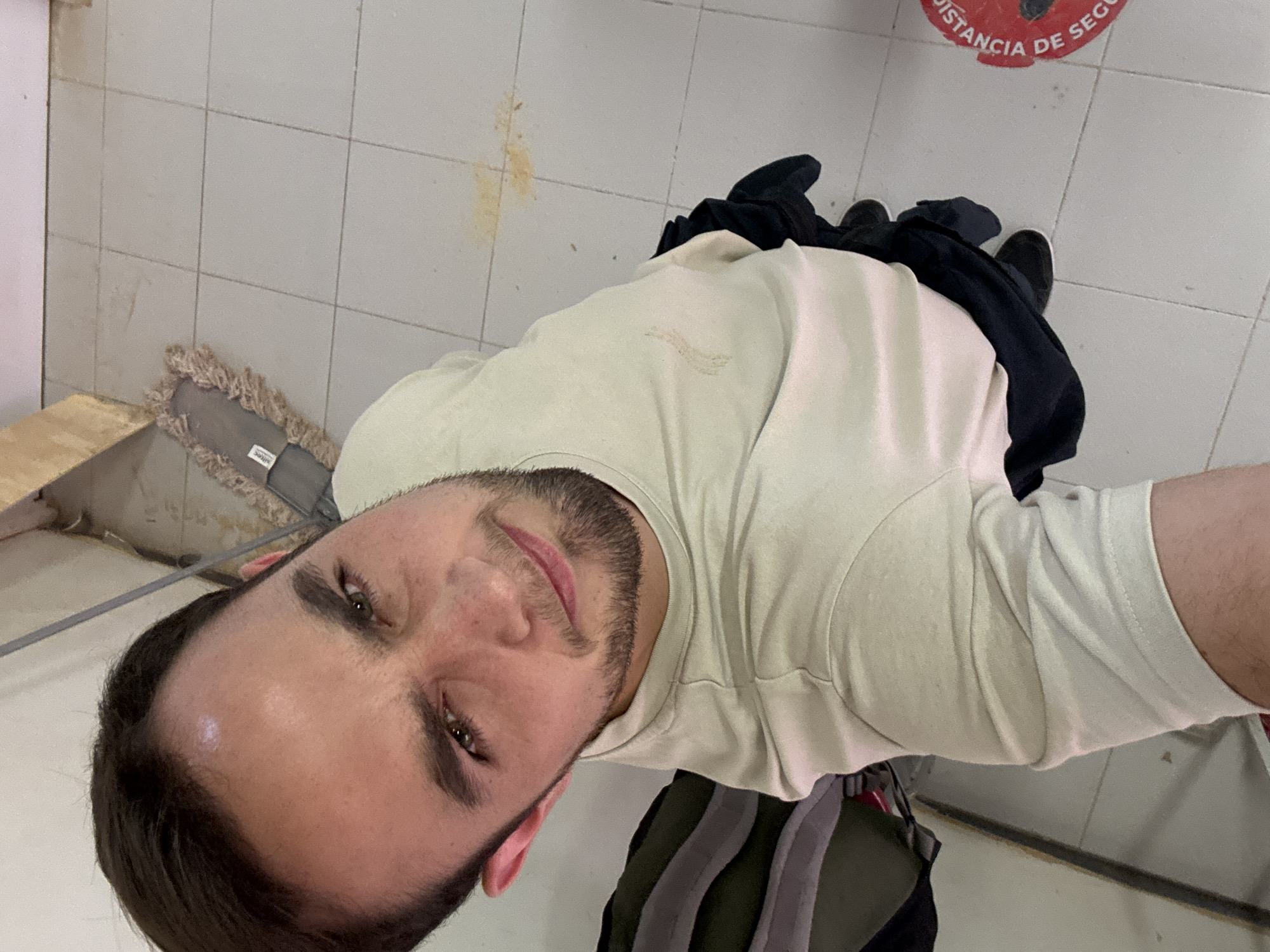 A medium shot of a man taking a selfie indoors. He is wearing a light-colored t-shirt and dark pants with black shoes. The background appears to be a tiled floor, possibly a restroom or utility area. A mop leans against the wall near the bottom left. There is a red circle on the floor with the Spanish words "DISTANCIA DE SEGURIDAD," which translates to "SAFETY DISTANCE." There are brown stains on the floor. The location cannot be verified, nor can it be determined to be San Bernardino Tlaxcalancingo, Mexico.