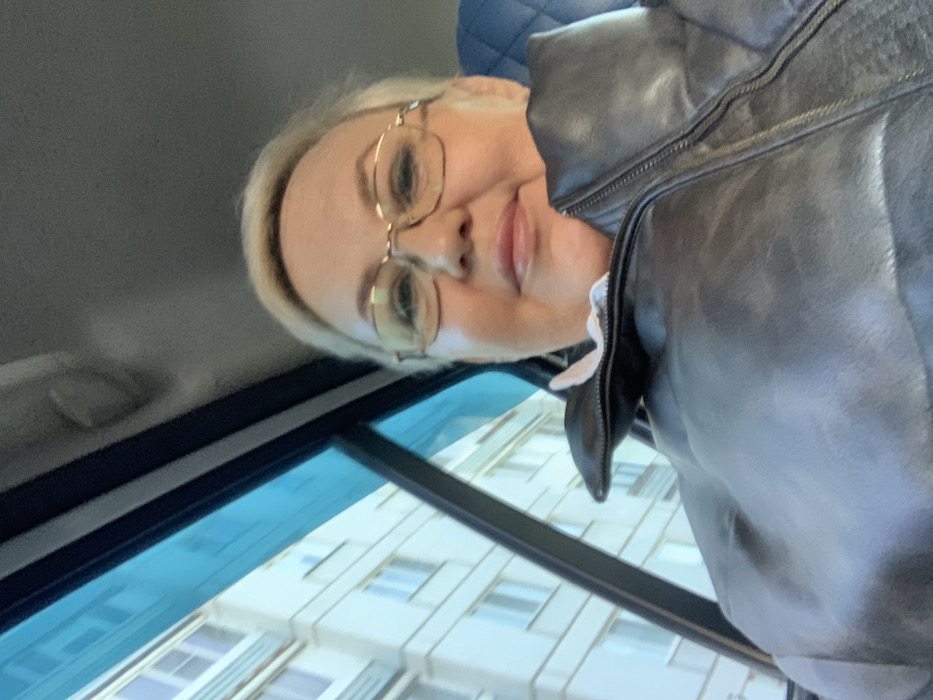 An adult female is depicted in a close-up selfie, seated within a vehicle in the city of Chaikovsky, Russia. She has short, light-colored hair and is wearing gold-framed prescription glasses. Her attire consists of a dark, zippered jacket, possibly leather, with a white collar visible underneath. The subject looks directly at the camera with a neutral expression. Through the vehicle's blue-tinted side window, a light-colored, multi-story residential building with numerous rectangular windows is visible in the background. Elements of the vehicle's interior, including a dark, diamond-stitched seat, are discernible behind her.
