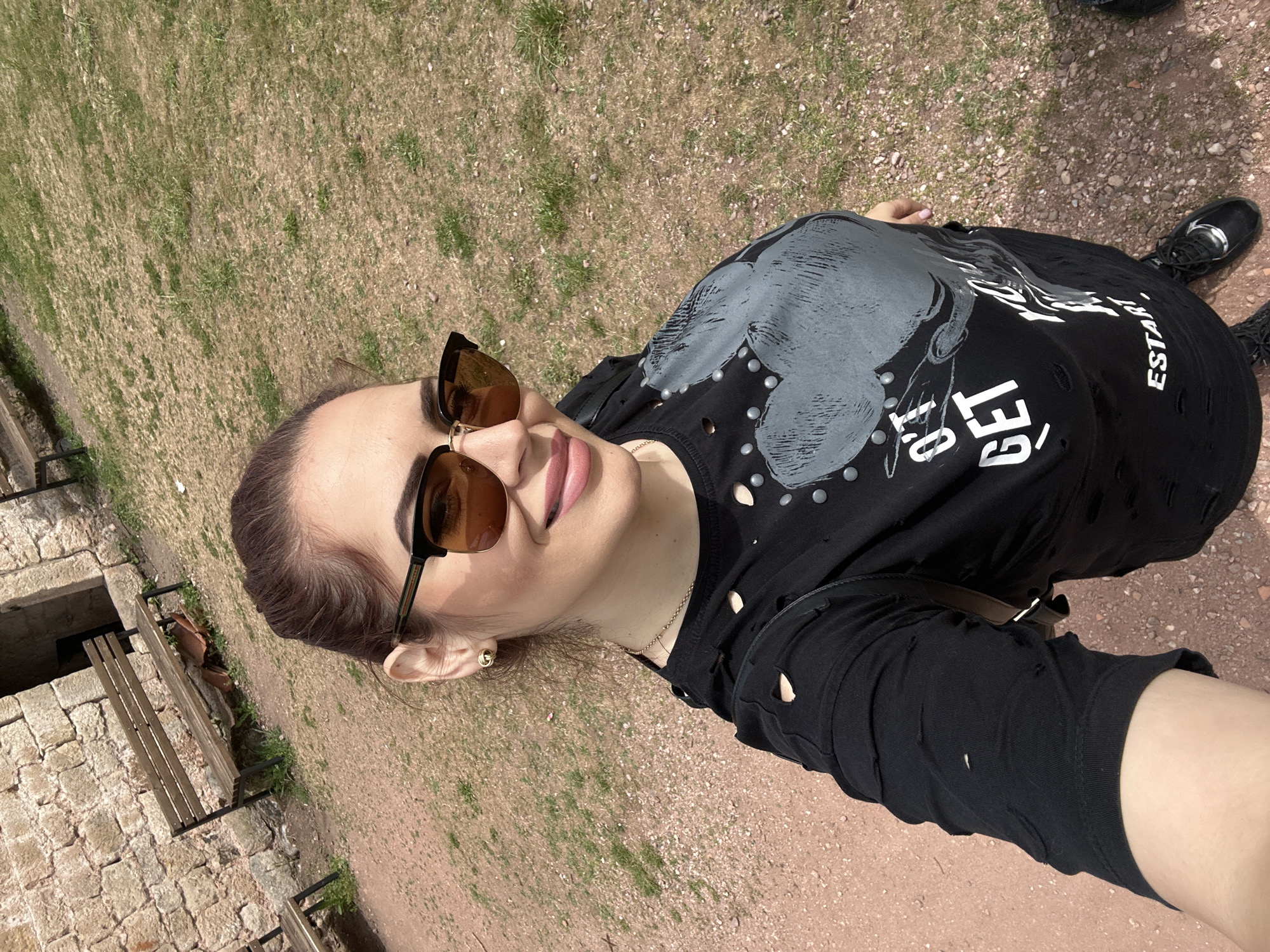 A woman stands outdoors in Belogradchik, Bulgaria. The ground beneath her is a mix of red soil, small pebbles, and dry grass. To the left, a partial view of a stone wall structure and a wooden bench are visible. The woman is wearing a black t-shirt with a graphic design, stud accents, and distressed cut-outs. She also wears dark sunglasses and a gold earring. Her hair is pulled back. In the background, a black shoe can be seen on the ground. The perspective suggests the photo was taken from a slightly elevated angle, possibly as a selfie.