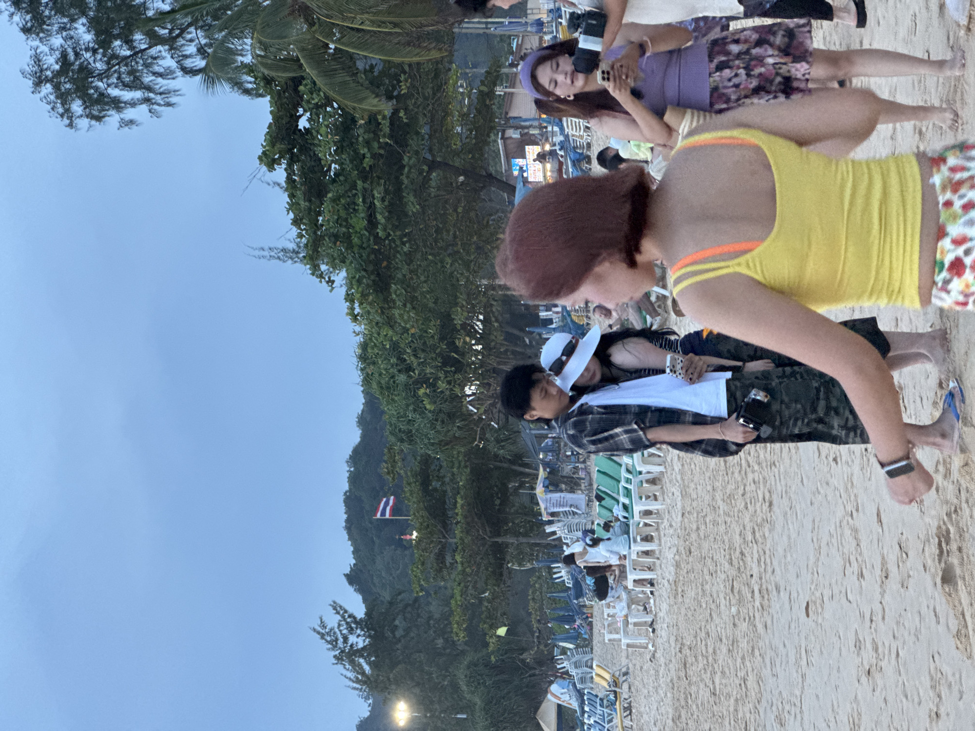 The image depicts a beach scene in Karon, Thailand, likely during twilight hours. In the foreground, a person with short reddish-brown hair wearing a yellow tank top with orange straps and a floral-patterned skirt is shown from behind, facing away from the camera. To their right, another person is visible, also from behind, wearing a purple tank top and a floral skirt, holding a smartphone and a small camera.
In the middle ground, a couple is standing close together. The male, on the left, is wearing a plaid shirt over a white t-shirt and camouflage pants, holding a black camera. The female, on the right, is wearing a white sun hat and a light-colored garment.
The background reveals a beach populated with numerous lounge chairs and umbrellas, some of which are set up in rows. Beyond the beach, lush green trees and vegetation cover a hilly landscape, with a Thai flag visible flying atop one of the hills. The sky is overcast with a pale blue hue, suggesting the end of the day. The overall impression is a relaxed, tourist-oriented beach environment.