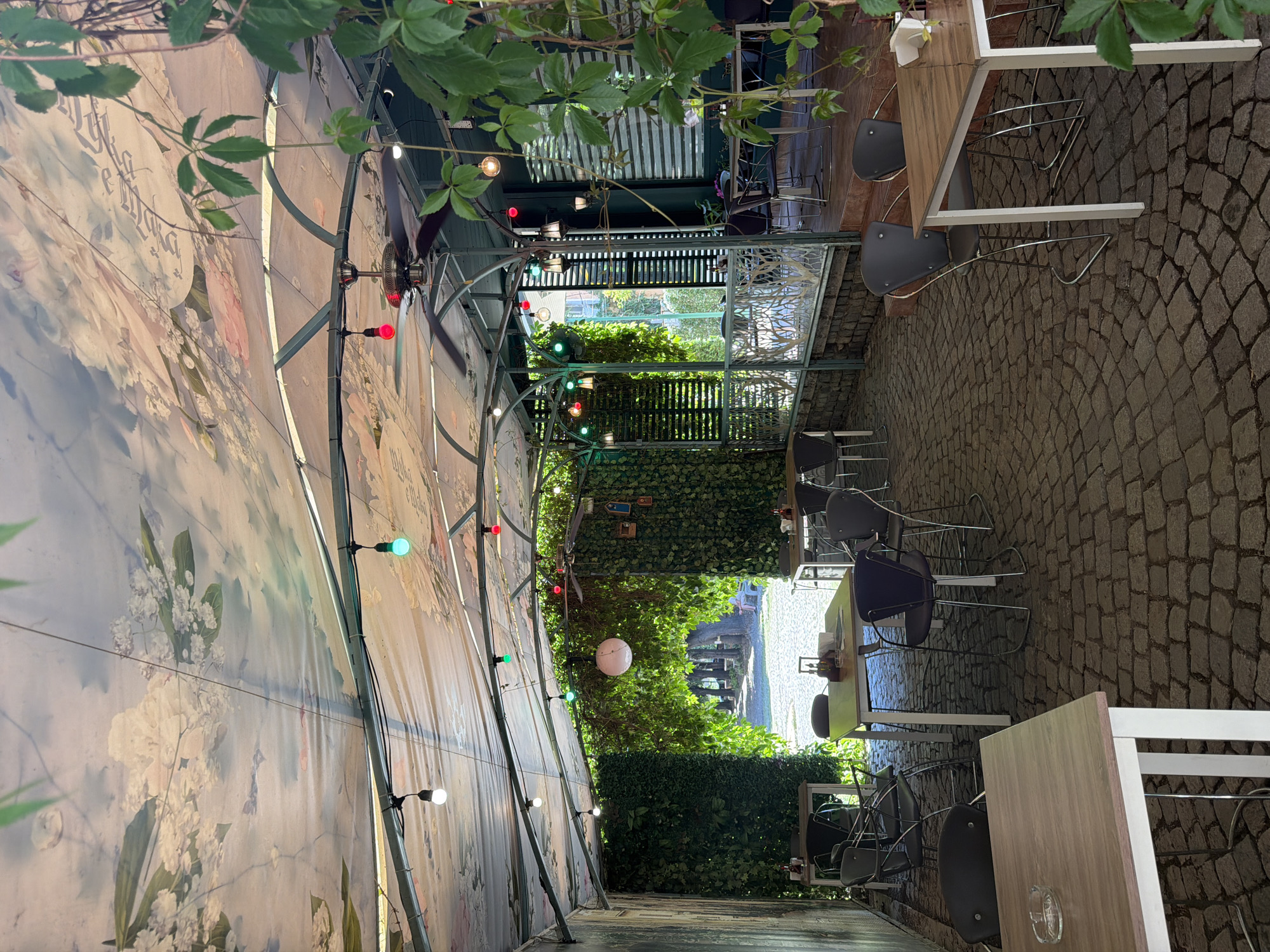 An outdoor seating area of a restaurant is pictured. The seating area is covered by a canvas canopy decorated with a floral mural. String lights are suspended from the canopy, with some illuminated in various colors. Several tables and chairs are arranged on a cobblestone ground. To the left, a wall is covered in ivy, and to the right, there is a building with windows covered by blinds. Greenery hangs from above and trails down the right side of the image. The scene appears to be in the city of Pazardzhik, Bulgaria.