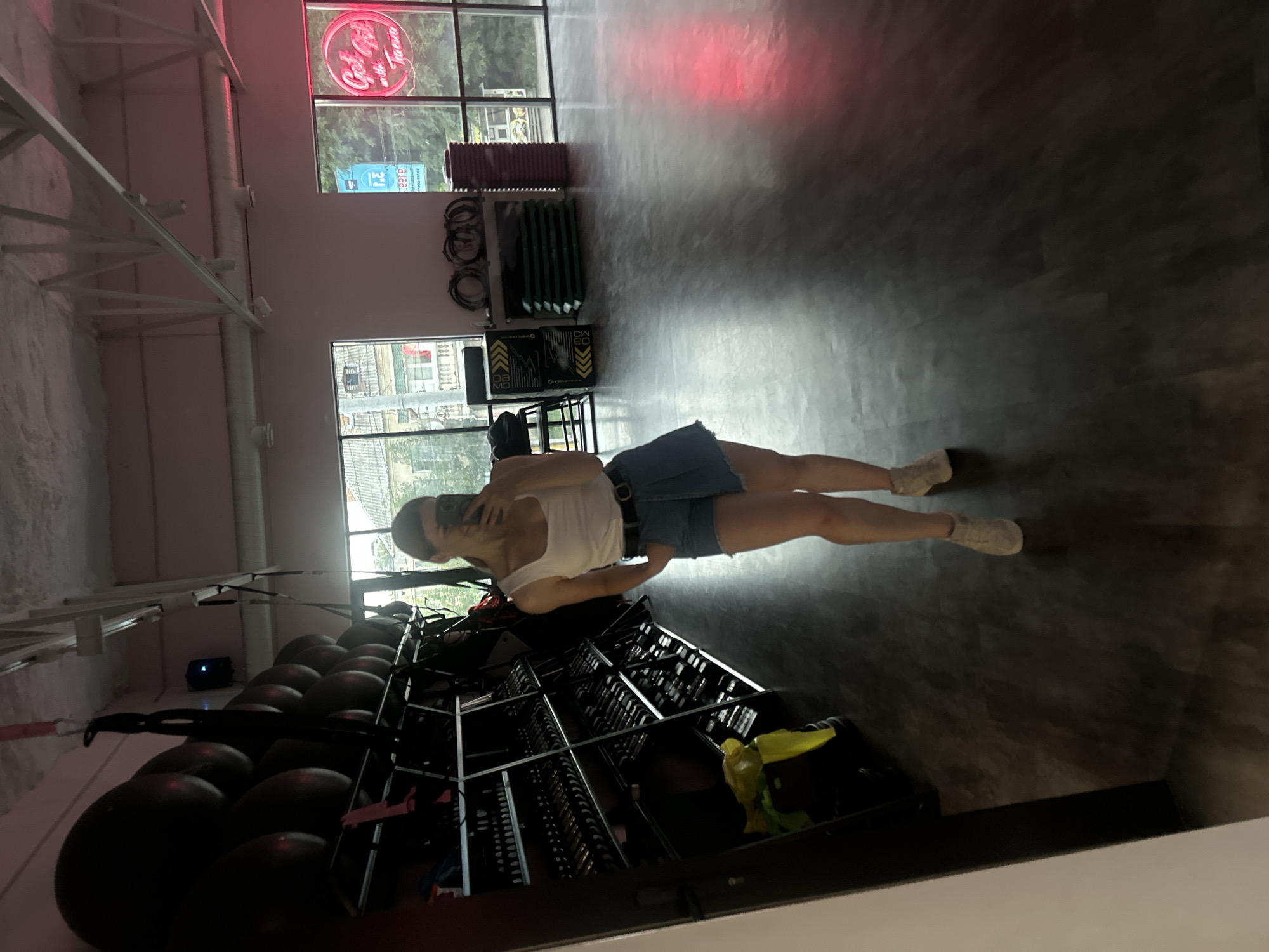 A full-body mirror selfie is taken by a young woman inside a gym in Ternopil, Ukraine. The woman, dressed in a white tank top, denim shorts, and white sneakers, stands with her hands in her pockets and holds a phone to take the picture. In the background, rows of gym equipment, including weights and resistance bands, are visible. Large windows overlook a street scene in Ternopil. A neon sign with the text "Got Girl" is prominently displayed on the wall. The floor of the gym is a dark, polished wood.