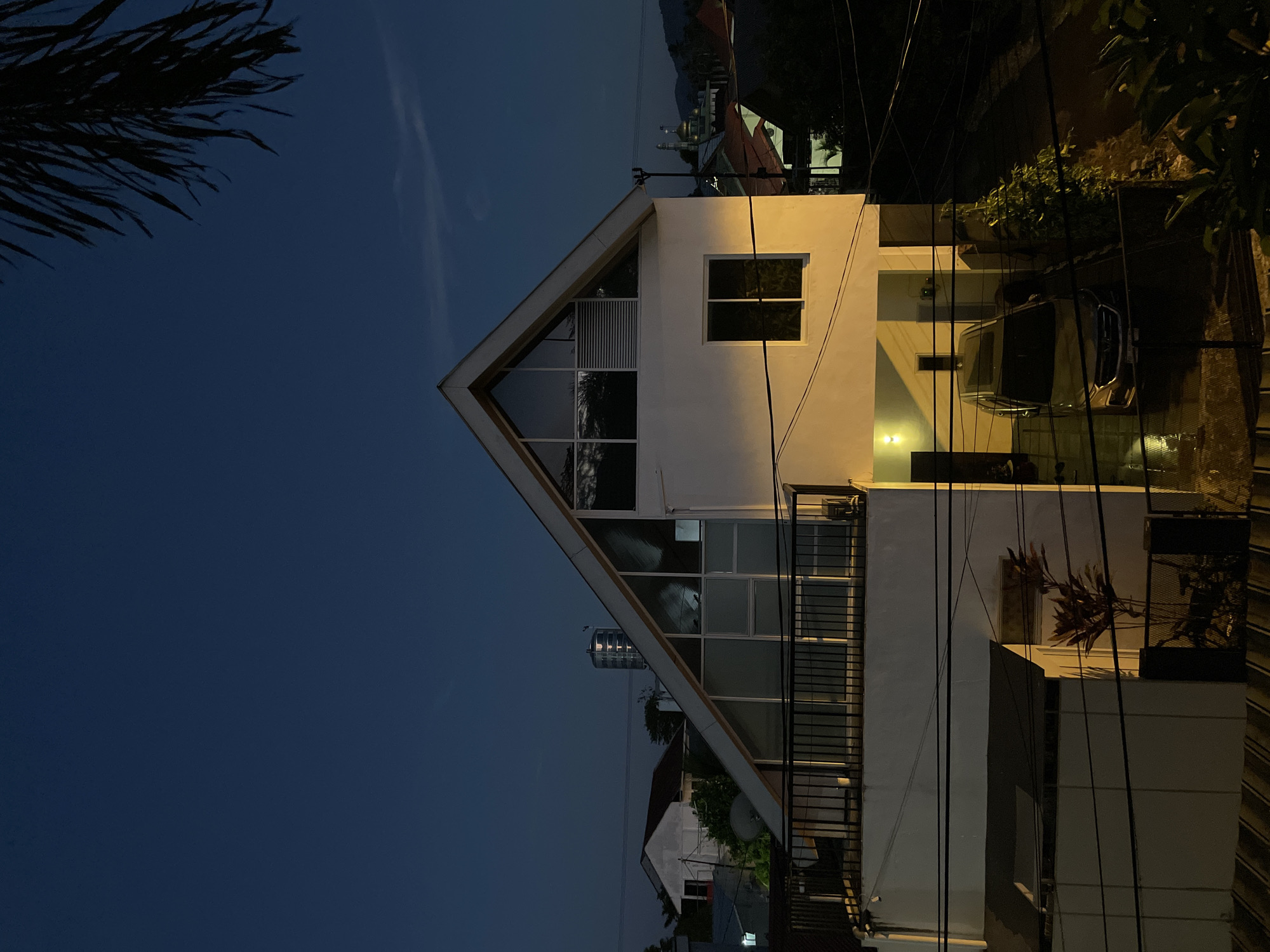 This image depicts a residential area at dusk in Manado, Indonesia. A modern, white house with a distinctive triangular roof dominates the foreground. The house features large, multi-paned windows on the upper floor and a single rectangular window on the lower floor, with a covered carport sheltering a silver sedan. A black metal railing encloses a balcony on the second level.
Surrounding the main house are other residences, some with white walls and red roofs, and a building with a prominent satellite dish. In the distance, a cylindrical tower structure is visible, along with the silhouette of a mosque with golden domes against the darkening sky. The scene is illuminated by artificial lights from the houses and streetlights, casting long shadows. Electrical wires crisscross the frame, typical of urban Indonesian environments. A palm frond hangs into the upper right corner of the image, adding a touch of tropical foliage. The sky is a deep, clear blue, indicating the transition from day to night. The ground beneath the carport appears wet, suggesting recent rain. No people are visible in the image.