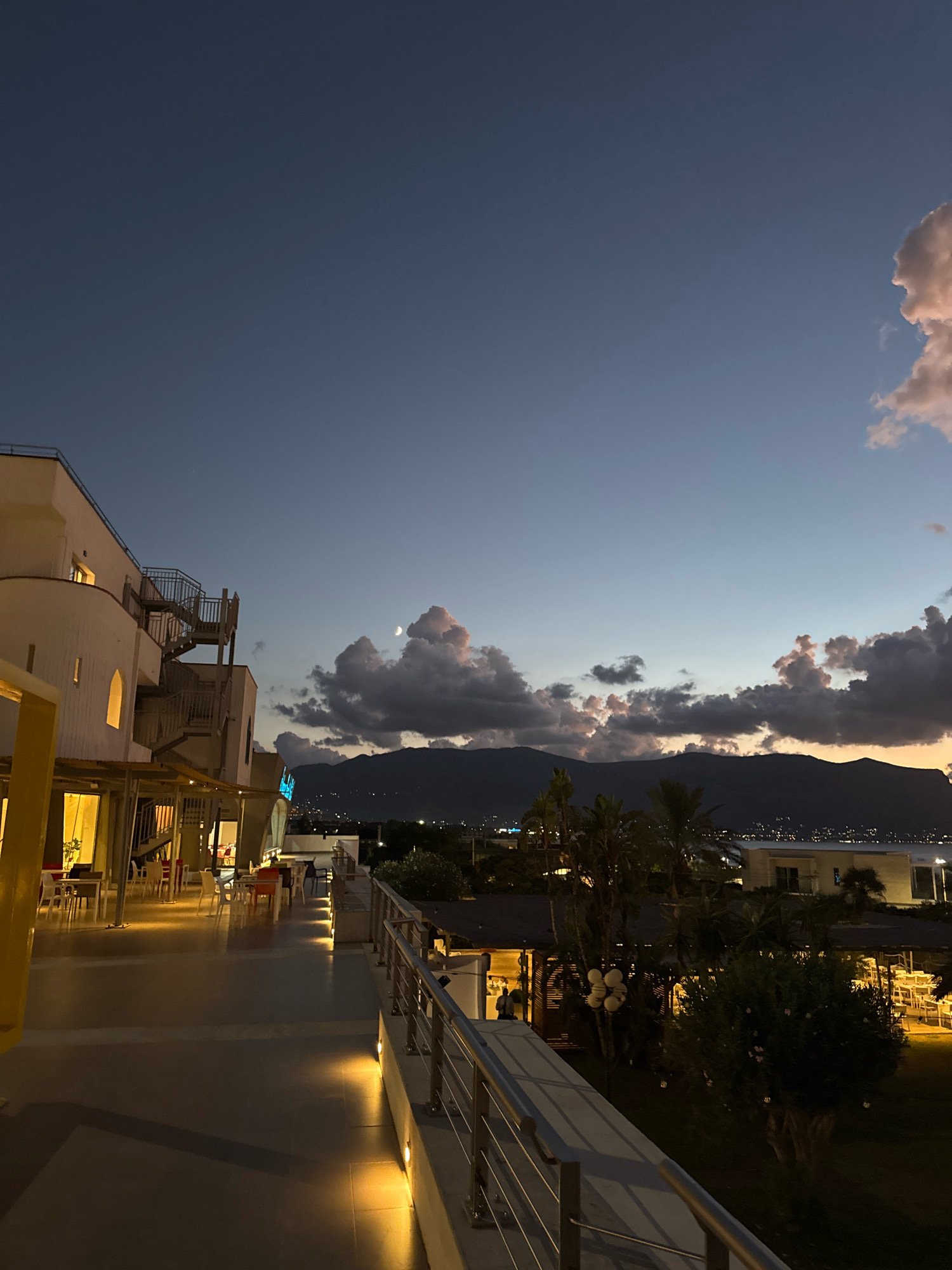 This is a vertical, eye-level shot of a restaurant terrace in Isola delle Femmine, Italy, taken at dusk. A multi-story building with an external staircase occupies the left side of the frame, with tables and chairs set up on its ground-level patio. The terrace railing extends to the right, overlooking a courtyard with more outdoor seating, trees, and illuminated buildings in the background. A sliver of moon and scattered, colorful clouds are visible in the darkening sky above a silhouetted mountain range and a distant city skyline.