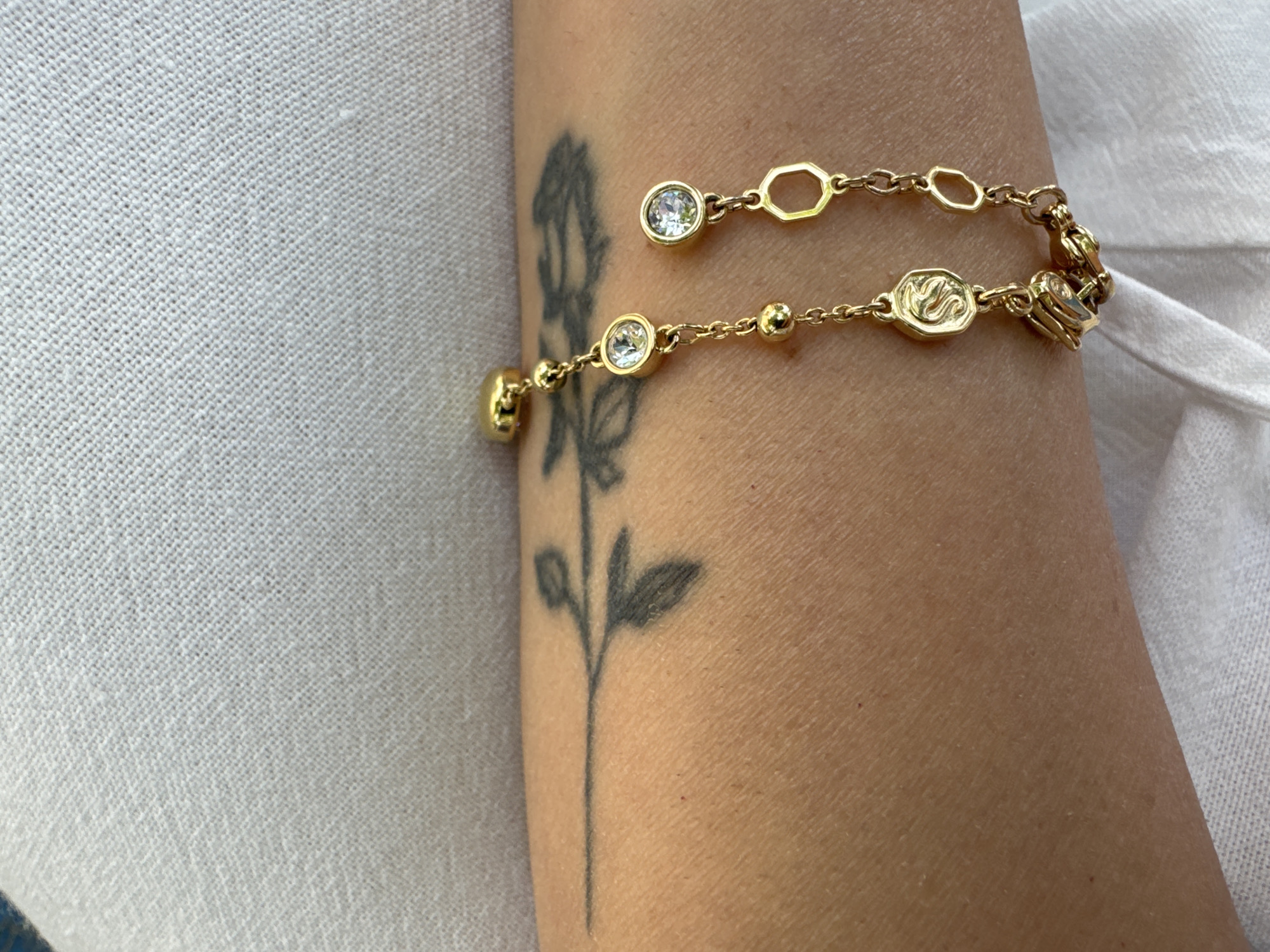The image shows a close-up view of a person's arm, displaying a tattoo of a thin-lined branch with a few leaves. A delicate gold bracelet with small, clear gemstones, beaded links, and a charm shaped like a stylized initial "S" is worn on the arm. The arm rests against a textured, light-colored fabric, possibly clothing or a surface. The background is out of focus and appears to be a continuation of the same textured fabric. The image is well-lit, highlighting the details of the bracelet and the tattoo. There is no identifiable location within the city of Sofia, Bulgaria.