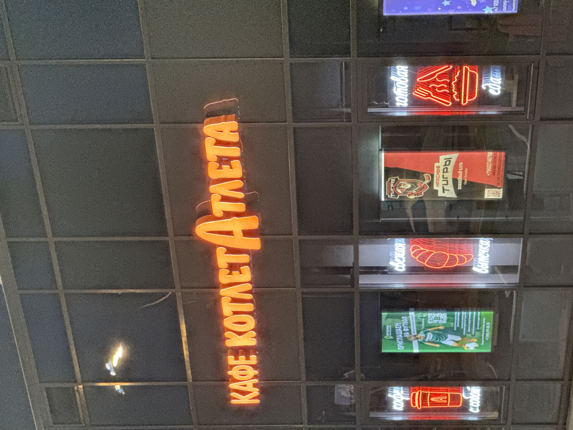 A neon sign above a storefront reads "КАФЕ КОТЛЕТ АТЛЕТА" in large, orange, backlit letters. Below the main sign, individual window displays feature neon signage and advertisements. On the left, a red neon coffee cup with the letter "A" inside is displayed, with the words "кофе с собой" (coffee to go) in white neon below. Next to it, a green advertisement displays a young man in a soccer uniform kicking a ball and reads "ПРИГЛАШАЕМ НА ФУТБОЛ" (We invite you to football). To the right of that, a red neon sign shows a croissant and the words "свежая выпечка" (fresh pastries) in white neon. Further to the right, a black and red advertisement for "КРАСНЫЕ ТИГРЫ" (Red Tigers) hockey club is visible. Next to that, a red neon sign depicts a steaming bowl of food with cutlery, and the words "готовая еда" (ready-made food) in white neon. The storefront appears to be located in Krasnoye Selo, Russia, given the presence of the "Red Tigers" hockey club advertisement, a team associated with the area. The overall scene depicts an evening exterior of a cafe or eatery, illuminated by neon signs.