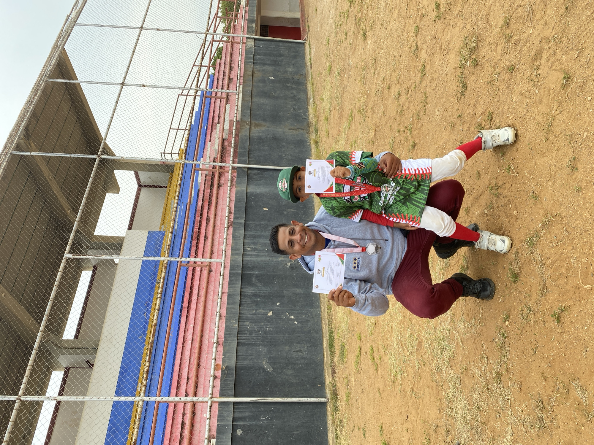 The image depicts a man and a young boy posing on a baseball field near a stadium, likely in Juan Griego, Venezuela. The man is kneeling and holding the boy, who is dressed in a green and white baseball uniform with red socks and a matching cap. Both are wearing medals around their necks and holding certificates, indicating they likely participated in a baseball event and received awards. The stadium behind them has seating painted in blue, yellow, pink, and white. The ground appears to be dirt and sparse vegetation. The lighting suggests it is daytime.