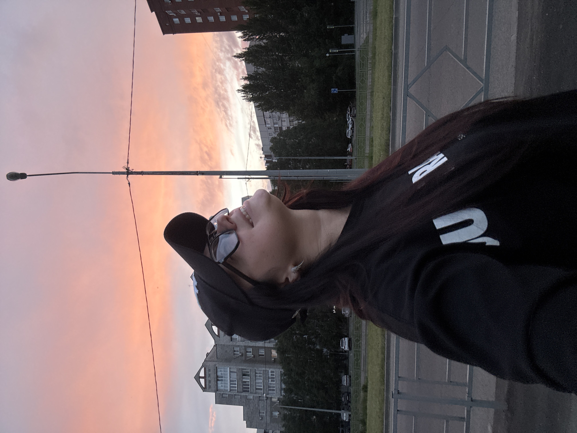 A female subject, wearing a black baseball cap and dark sunglasses, is positioned in the left foreground, looking upwards with a slight smile. Her long, dark hair cascades over her right shoulder. She wears a dark long-sleeved top with white lettering, partially displaying "JU" and "R". A silver-toned, spiky earring is visible on her right ear.
The background depicts an urban environment under a sky transitioning from pink and orange hues at the horizon to lighter grey-blue overhead, indicative of dawn or dusk. A tall, dark streetlamp with an overhead light fixture dominates the central background, with two parallel wires extending horizontally across the frame.
To the left, a multi-story grey concrete building with distinctive gabled roof sections and numerous windows is visible. To the far right, a taller, multi-story building with a reddish-brown facade and multiple windows, some with air conditioning units, stands. A line of dark green deciduous trees occupies the middle ground.
A metal guardrail with a diamond pattern is present in the lower foreground, separating a grassy area from a paved road. Several light-colored cars are parked in lots beyond the trees. A blue rectangular street sign is visible on the right side of the road. The location is an unknown city, Unknown.
