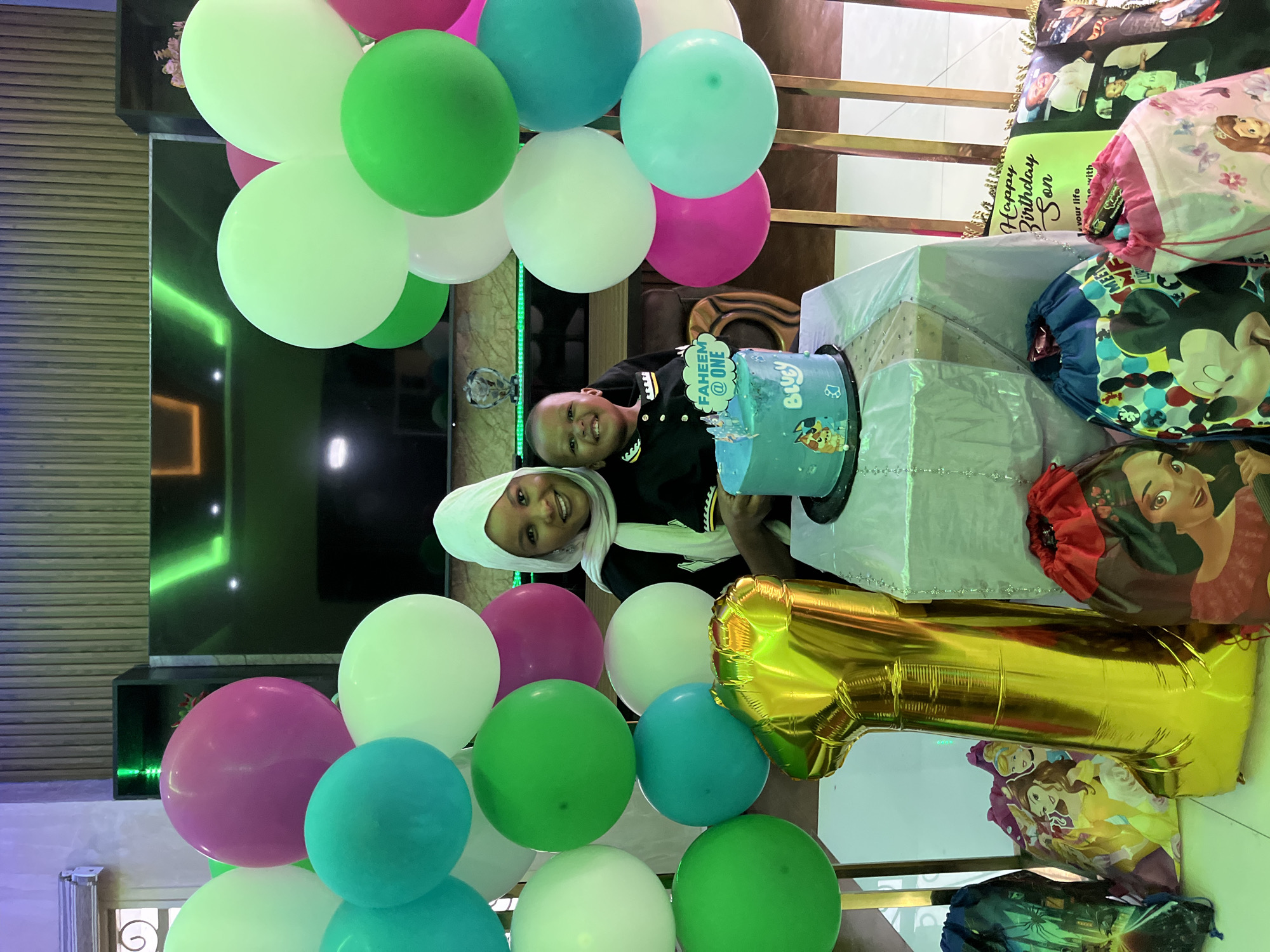 A woman and a young child are posing for a first birthday celebration in an indoor setting in Jos, Nigeria. The woman, with light brown skin, is smiling and wears a white hijab and a black collared top with white and yellow accents on the placket. She is holding a smiling dark-skinned male child, who appears to be one year old, dressed in a black collared shirt with a patterned design.
In the center, on a table draped with a shimmering silver fabric, sits a two-tiered light blue birthday cake with a "Bluey" theme. A cake topper reads "FAHEEM @ ONE." Surrounding the subjects are numerous balloons in clusters, featuring colors such as vibrant pink, various shades of green, white, and teal. These balloons frame the scene on both the left and right sides, some supported by metallic, gold-colored vertical stands.
On the light-colored floor in the foreground, a large gold metallic foil balloon shaped like the number "1" rests. Beside it and to the right are several drawstring gift bags, each featuring animated characters: Disney princesses (Belle, Cinderella), Elena of Avalor, and Mickey Mouse, along with a pink bag showing another princess. Behind these bags, a light green pillow or soft banner with "Happy Birthday Son" written in script and several printed photographs is visible.
The background shows a modern interior with vertical wooden wall paneling in the upper section and a large dark flat-screen television reflecting distinct green ambient lighting. To the right of the television, a dark shelving unit also displays green accent lighting. The overall environment suggests a decorated living space or an event hall.