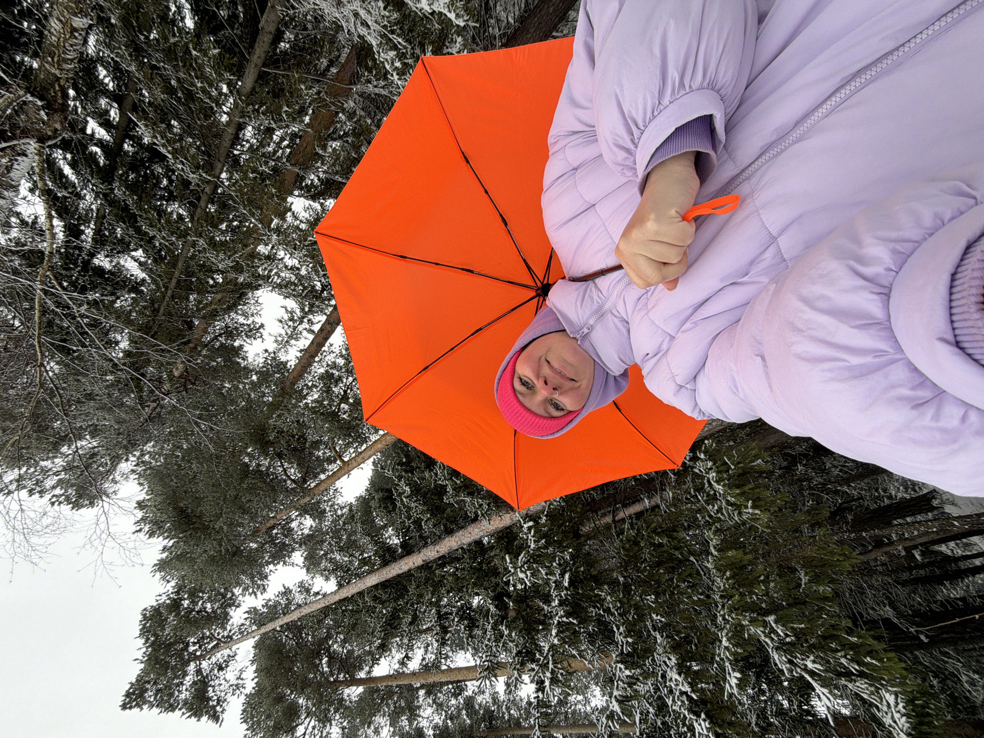 The image shows a person holding an open orange umbrella. The person is wearing a pink beanie and a lavender puffer jacket. Trees with snow on their branches are visible in the background. The location is Chaikovsky, Russia.