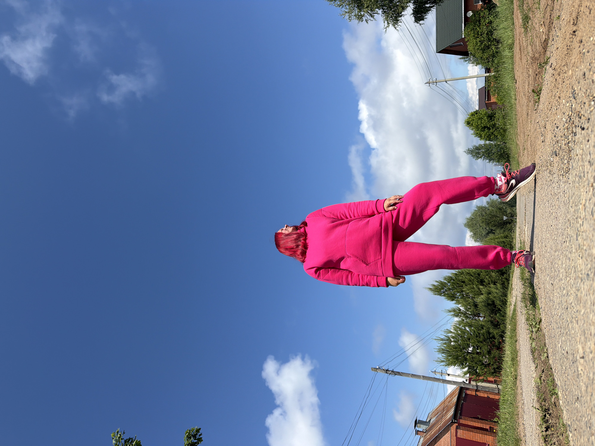 The image, correctly oriented, depicts a person standing on a dirt path under a vast, bright blue sky with scattered white clouds. The subject is a woman with vibrant, bright pink or red hair, dressed in a matching bright pink tracksuit and purple and pink athletic shoes. She stands with her back partially to the viewer, looking upwards towards the sky. Her left arm is slightly bent, and her right arm hangs alongside her body.
The setting is an outdoor, semi-rural environment, consistent with a village or countryside road in Chaikovsky, Russia. The foreground shows a light-colored dirt or gravel road. To the right of the road, there is a grassy verge and a dense line of green trees and bushes, including some conifer-like trees.
In the mid-ground, several wooden utility poles with power lines are visible, stretching across the scene. Further in the background, two houses can be seen: one with a dark green roof partially obscured by trees, and another brick-built structure with a reddish, possibly corrugated metal roof.
The time of day appears to be bright daylight, either morning or afternoon, given the clear illumination and the quality of the sunlight on the clouds. There are no discernible texts or specific emotions visible on the person's face due to her pose.