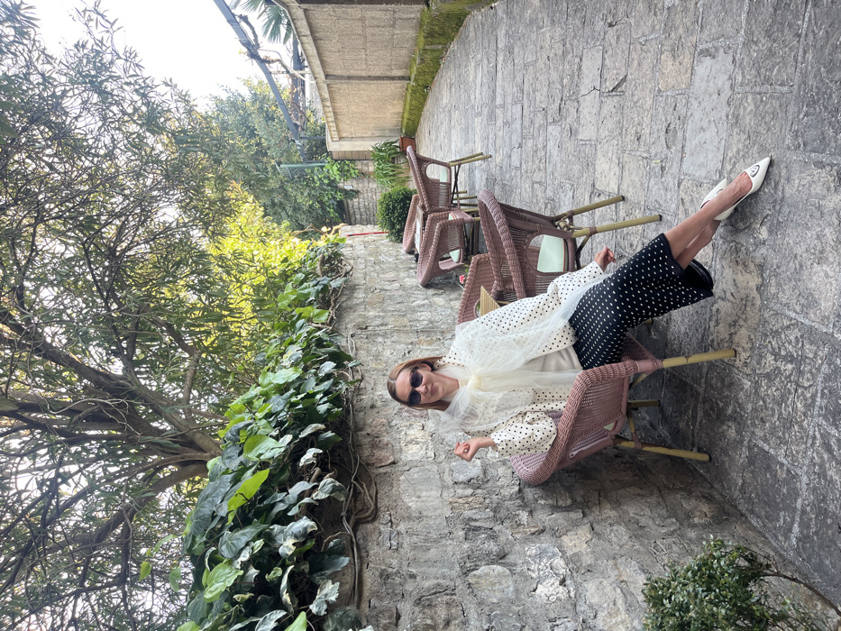 The image depicts an outdoor cafe or patio situated within a narrow, stone-paved passage in the Old Town of Budva, Montenegro. A single woman with reddish-brown hair, wearing sunglasses, is seated in a light brown wicker-style chair with a light green cushion, positioned against a rough-hewn stone wall largely covered by green ivy. She is dressed in a white long-sleeved top with black polka dots, a white sheer scarf tied around her neck, a light-colored top underneath, black culottes or a midi-skirt with white polka dots, and white pointed-toe heels featuring a dark embellishment. Her right hand rests on the chair's armrest, and she exhibits a slight smile while looking slightly off-camera to her right.
To the right of the woman, a series of identical empty wicker chairs with light green cushions are arranged around small square tables, extending down the stone-paved pathway. The pathway is flanked on the right by a textured, light beige wall with moss growth at its base and several potted plants. Overhead, a dense canopy of tree branches with green foliage provides natural cover, with additional trees and greenery visible in the background against a bright, clear sky. The scene is well-lit by natural daylight.