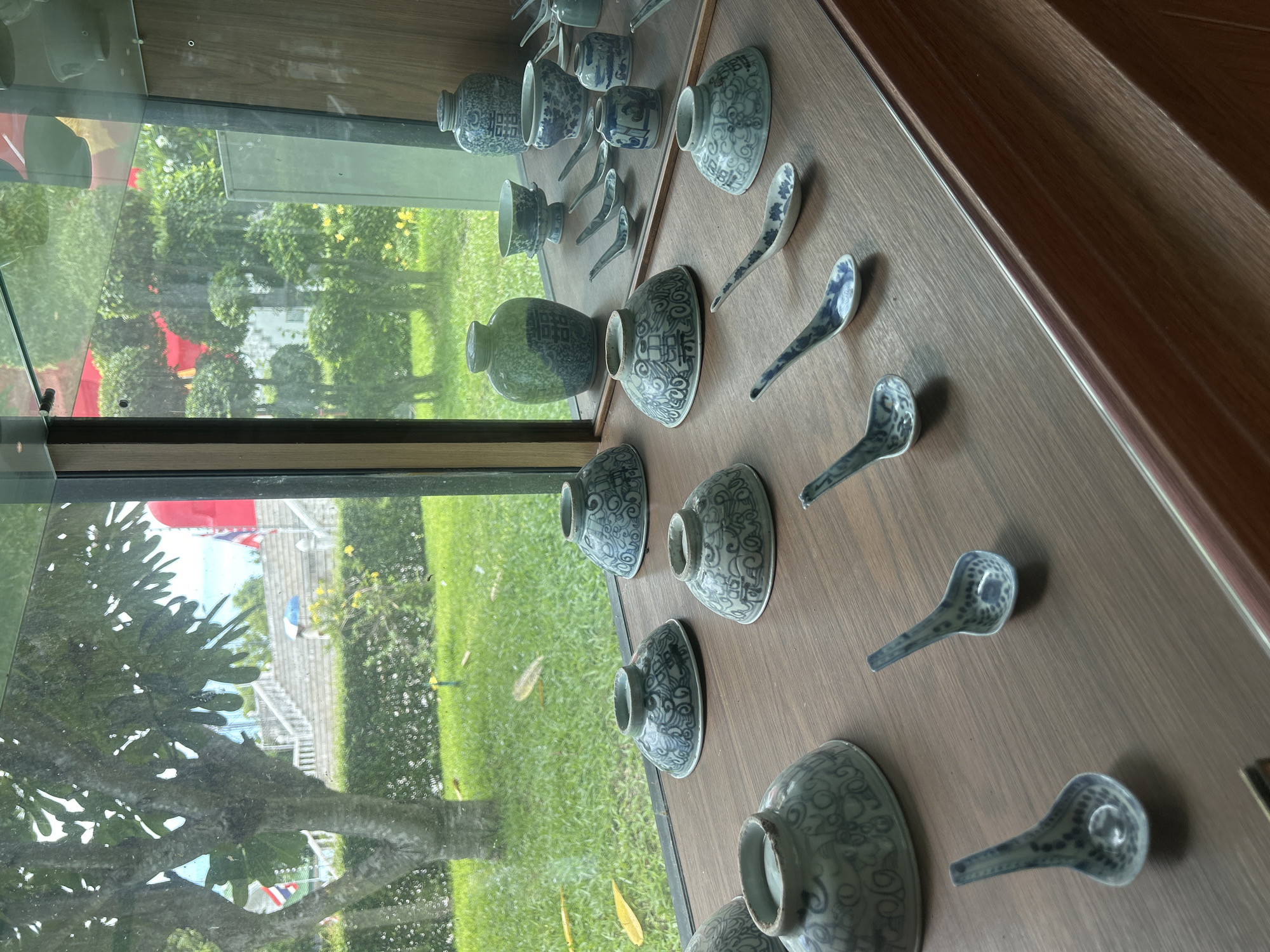 The image displays a collection of ceramic bowls and spoons arranged on a wooden shelf inside a glass display case. The ceramics are predominantly blue and white with intricate traditional patterns. In the background, visible through the glass, is a lush green lawn with trees and a body of water, suggesting an outdoor environment. Further in the background, there is a white staircase leading upwards, with flags and red structures visible, indicating a public or ceremonial space. The scene appears to be an exhibition of decorative ceramics, possibly located in a museum or cultural center in Thai Ko Subdistrict, Thailand, given the visual cues of tropical vegetation and what could be national flags. There are no people visible in the image.