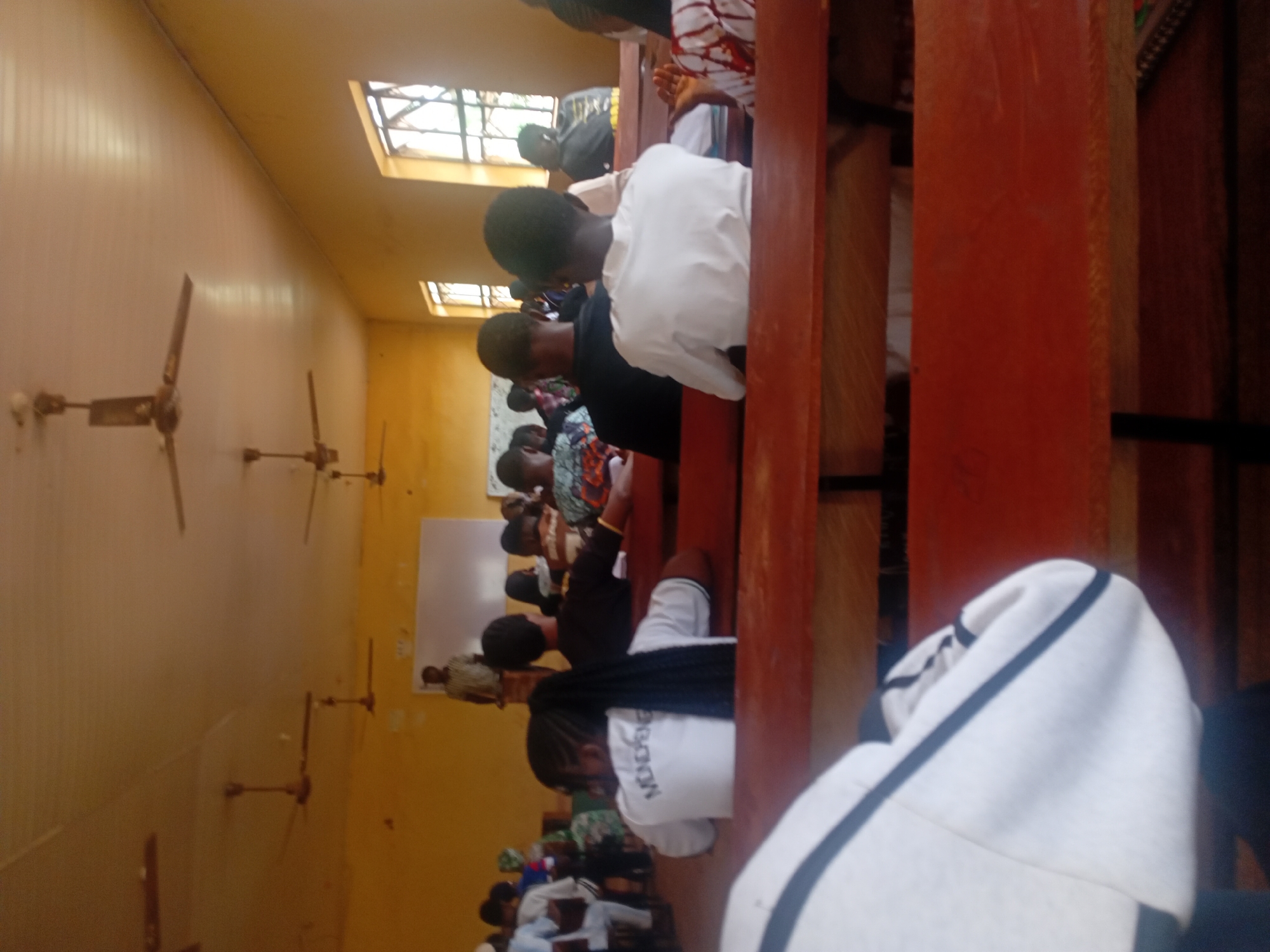 The image shows a gathering of people inside a room, likely a classroom or meeting space. The location appears to be in a building in the city of Unknown, Unknown. People are seated in rows facing what looks like a whiteboard. Most people are facing forward, seemingly engaged in a presentation or lecture. There are visible ceiling fans attached to the walls. The room is painted in a yellowish-cream color with natural light entering through high windows.