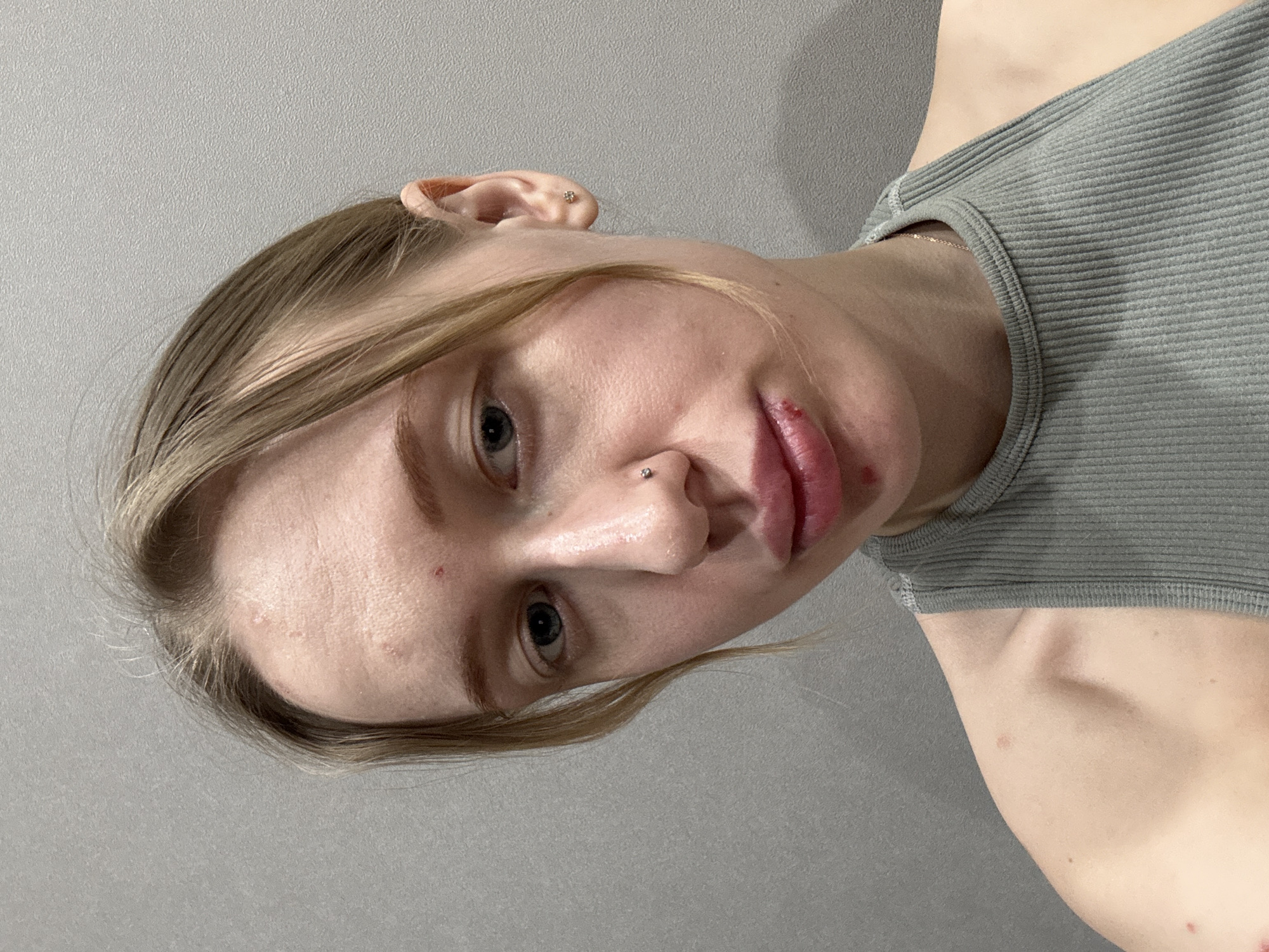 The image shows a close-up of a fair-skinned person with blond hair and blue eyes. The person is wearing a gray ribbed tank top. They have a nose piercing and ear piercings. The person has a visible blemish or sore on their lip and acne on their forehead and cheek. The background is a plain gray wall. There is no indication of the location being Rostov-on-Don, Russia, or any specific city.