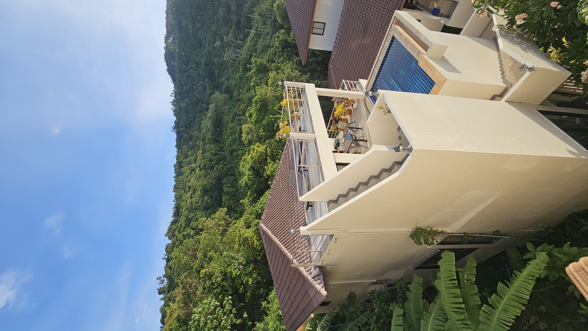The image shows a multi-story building with a swimming pool on the roof, surrounded by lush green vegetation and a hillside. The building has a neutral-colored exterior and a brown tiled roof. There are outdoor seating areas on multiple levels, including a rooftop terrace with a table and chairs, and a smaller balcony with potted plants. A blue swimming pool with a tiled bottom is visible on one of the upper levels. The background consists of a dense forest covering a hilly terrain, with a clear blue sky above. The scene appears to be a resort or a private residence in a tropical location. It is daytime, with sunlight illuminating the buildings and the surrounding landscape.