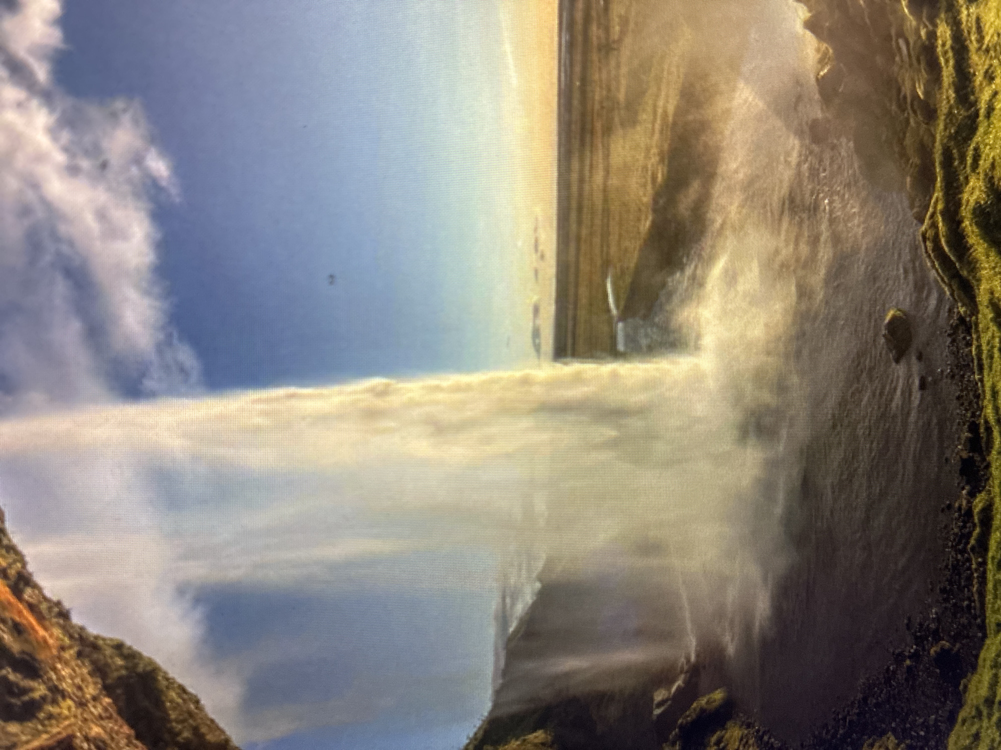 The image displays a majestic waterfall cascading down a rugged cliff face under a bright, partly cloudy sky. The scene is oriented vertically, with the powerful stream of white water and mist dominating the central view.
The waterfall plunges from a high ledge, creating a thick curtain of water and a significant amount of spray and mist, especially at its base. Sunlight strongly illuminates the falling water and mist, giving it a golden hue against the backdrop of the blue sky. To the left, a steep, rocky mountain slope with patches of brown and green vegetation is partially visible. On the right, a darker, moss-covered cliff with prominent rocky textures borders the waterfall. The ground where the water collects appears to be a rocky bed.
The sky above is clear blue, featuring large, white cumulus clouds on the left side and more diffused clouds towards the upper right. The bright lighting suggests it is daytime, likely a sunny day. No people or artificial objects are clearly discernible in the image, emphasizing the natural beauty and power of the landscape. There is no visible text. The image exhibits a pixelated grid pattern, suggesting it is a photograph taken of a digital screen. The location context provided is Asenovgrad, Bulgaria.