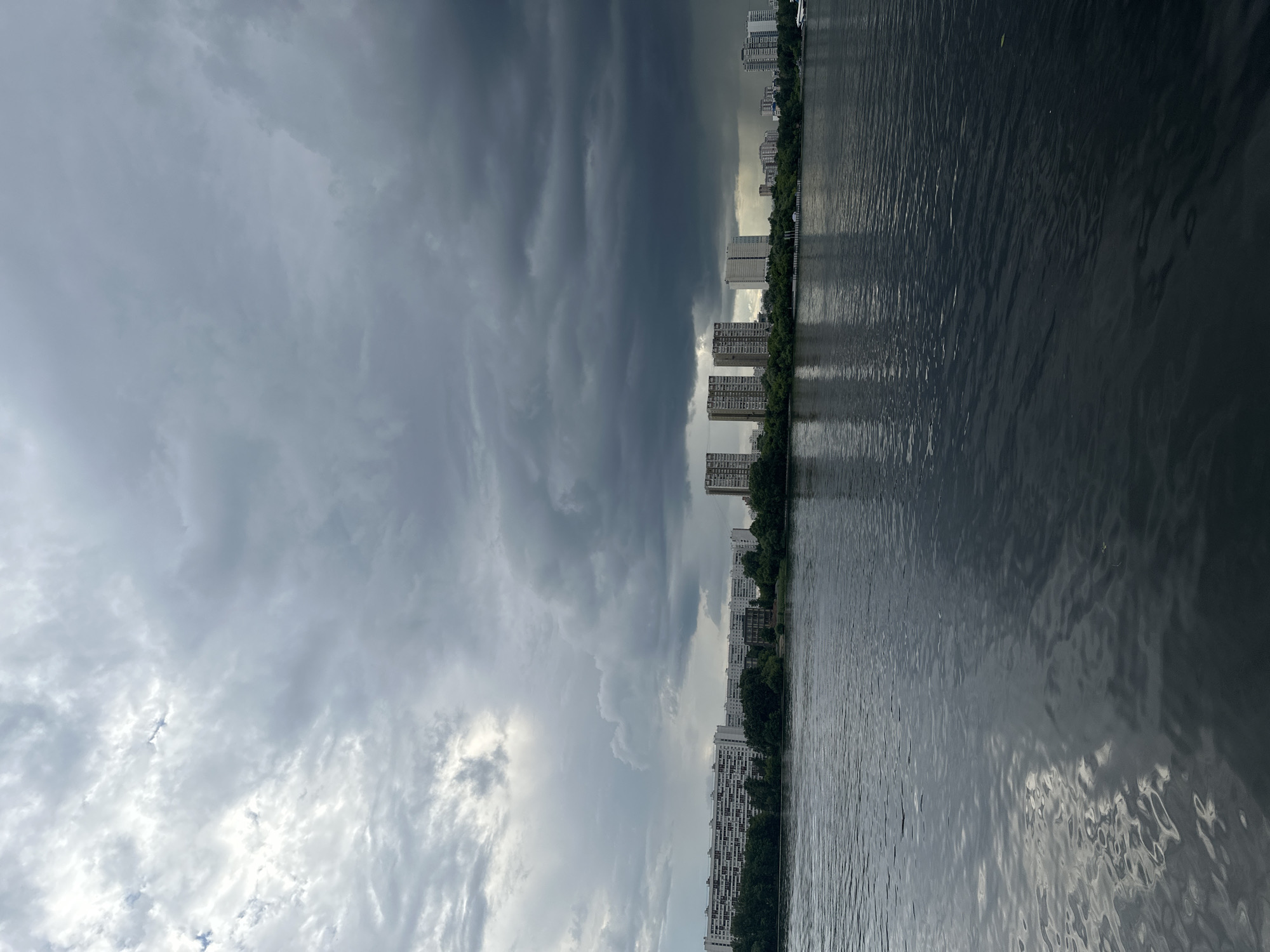 This is a vertical, eye-level shot of a cityscape on a cloudy day, viewed from a body of water. The scene is divided by the horizon line, with the sky occupying the left half of the frame and the water on the right.
The sky is dramatic, with a large expanse of dark, heavy clouds dominating the left side, suggesting an approaching storm or recent rain. Towards the right of the sky, there are patches of lighter clouds and hints of sunlight breaking through.
Along the distant shore, a line of white, multi-story apartment buildings is visible, interspersed with trees. These buildings create a clear urban silhouette against the sky. The water in the foreground is dark and rippled, reflecting the subdued light from the sky and the buildings. The reflections are distorted by the movement of the water, creating an abstract pattern.
There are no people or specific objects clearly identifiable in the scene. The overall mood is somewhat somber and dramatic due to the heavy clouds, contrasting with the more orderly, man-made structures of the city. The time of day appears to be late afternoon or early evening, judging by the quality of light. No visible text is present in the image.