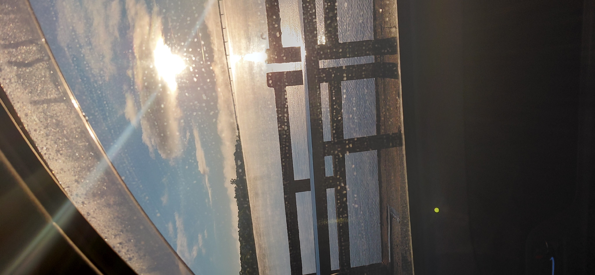 The image shows a view through a car window of a body of water, likely a river or lake, with a bridge in the distance. The sun is low in the sky, casting a bright glare on the water and creating lens flare effects on the window, which is also speckled with water droplets. In the foreground, a dark railing with rectangular openings is visible, partially obscuring the view. Mist or fog hangs low over the water, and the far bank of the water features a treeline. The sky is blue with scattered clouds. The image appears to have been taken from a moving vehicle. Without additional context, the specific location within Encarnacion, Paraguay, cannot be precisely identified.