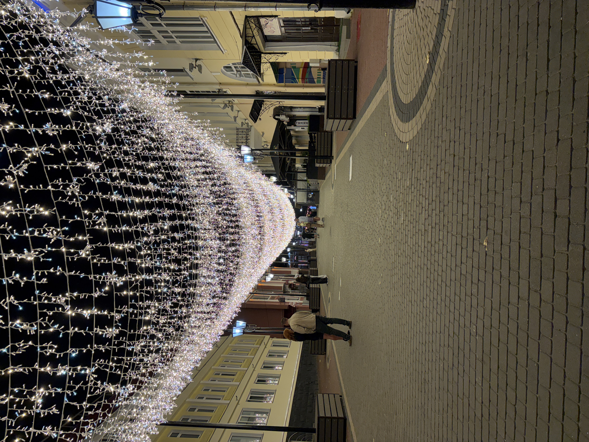The image depicts a pedestrian street in Vitsebsk, Belarus, illuminated by an overhead canopy of festive string lights. The street is paved with interlocking bricks, and a circular patterned inlay is visible on the right side of the pavement. Buildings with yellow facades line the street, featuring windows and awnings. Several people are present, including a couple standing in the mid-ground, facing each other. Other individuals are scattered further down the street, some appearing to be walking. Streetlights are strategically placed along the sidewalk. Planters containing foliage are visible at the base of some buildings. The overall scene suggests a nighttime setting, likely during a holiday season or a special event, given the extensive light decorations.