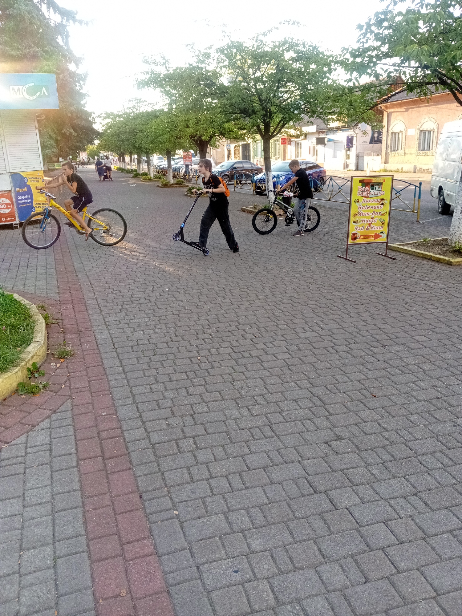 In the city of Tiachiv, Ukraine, on a paved sidewalk constructed of gray and red interlocking stones, three young individuals are engaged in recreational activities. The scene captures a sunny day, with trees lining the background and buildings visible.
On the left, a boy on a yellow bicycle is captured mid-motion, appearing to be either riding or dismounting. He is wearing a dark t-shirt and shorts. To his right, a second boy is operating a dark-colored scooter, leaning forward as if pushing off. He is dressed in a dark t-shirt and pants, with an orange backpack. In the background, a third boy is positioned near a black bicycle, appearing to be observing or waiting. He is also wearing dark clothing.
In the background, several cars are parked along the street. A white van is parked on the right side of the frame. A brightly colored yellow advertising sign, featuring text and images of food items, stands on the right side of the sidewalk. A white kiosk-like structure is visible on the far left, with blue signage. The overall environment suggests a public area, likely a promenade or pedestrian walkway. The light indicates it is either morning or late afternoon.