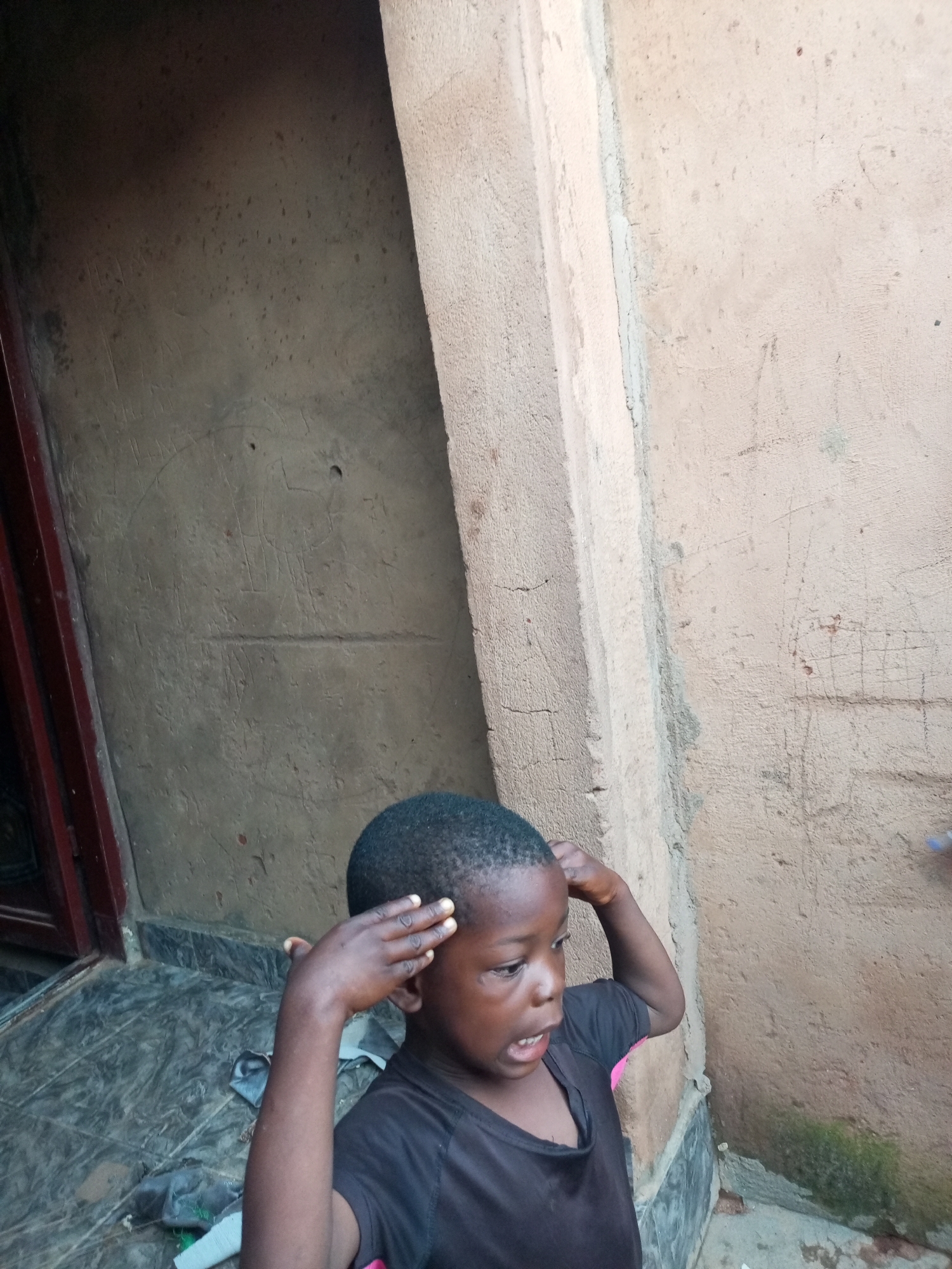 A child stands in front of a building with rough, textured walls, appearing to be in Tempure, Nigeria. The child, with dark skin and short black hair, is wearing a black t-shirt with pink accents on the sleeves. They are looking to their right with an open mouth and have their right hand raised to their forehead, as if in thought or greeting. The building's exterior is made of a rough, tan-colored material, with visible cracks and marks. A doorway with a dark red frame is visible to the left of the child, leading to an interior that is dark. The ground in front of the building is tiled with a dark grey, patterned material. Some debris, including dark fabric and a white object, is scattered on the ground.