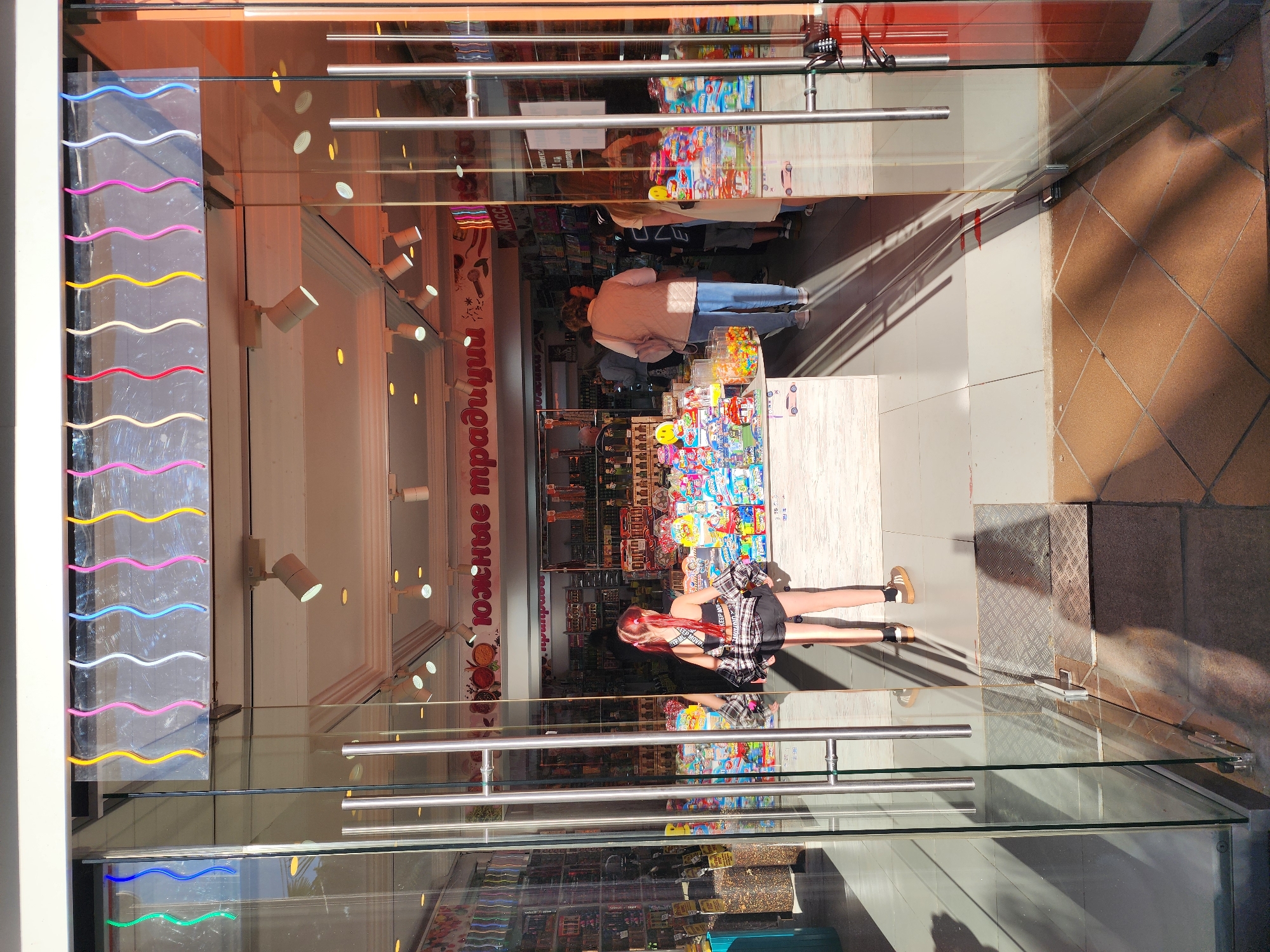 The image depicts the glass entrance of a shop, likely in Sochi, Russia, viewed from an external perspective on a bright, sunny day. Strong shadows are cast on the light brown tiled ground outside the entrance, indicating clear weather.
Through the glass, the interior of a brightly lit shop is visible, filled with colorful merchandise. A person with reddish hair, wearing a black top, a black and white plaid skirt, and light-colored sneakers, stands closer to the glass, seemingly looking into or through the shop. Further inside, another person wearing a light-colored jacket/vest and blue jeans is seen near a display counter.
The shop's interior features multiple shelves stocked with a variety of items, including candies, packaged goods, and toys. Spotlights are mounted on the interior walls, illuminating the displays. A prominent red sign inside the shop displays the Cyrillic text "Южные традиции" (Yuzhnye traditsii), which translates to "Southern Traditions."
On the left side of the glass entrance, a vertical decorative panel features a series of wavy, neon-like lights in various colors, including blue, white, pink, yellow, and red. The glass door has horizontal metal handles. Reflections on the glass create a layered view, merging the shop's interior with subtle hints of the external environment. A textured metal plate is visible on the ground directly outside the shop's threshold.
