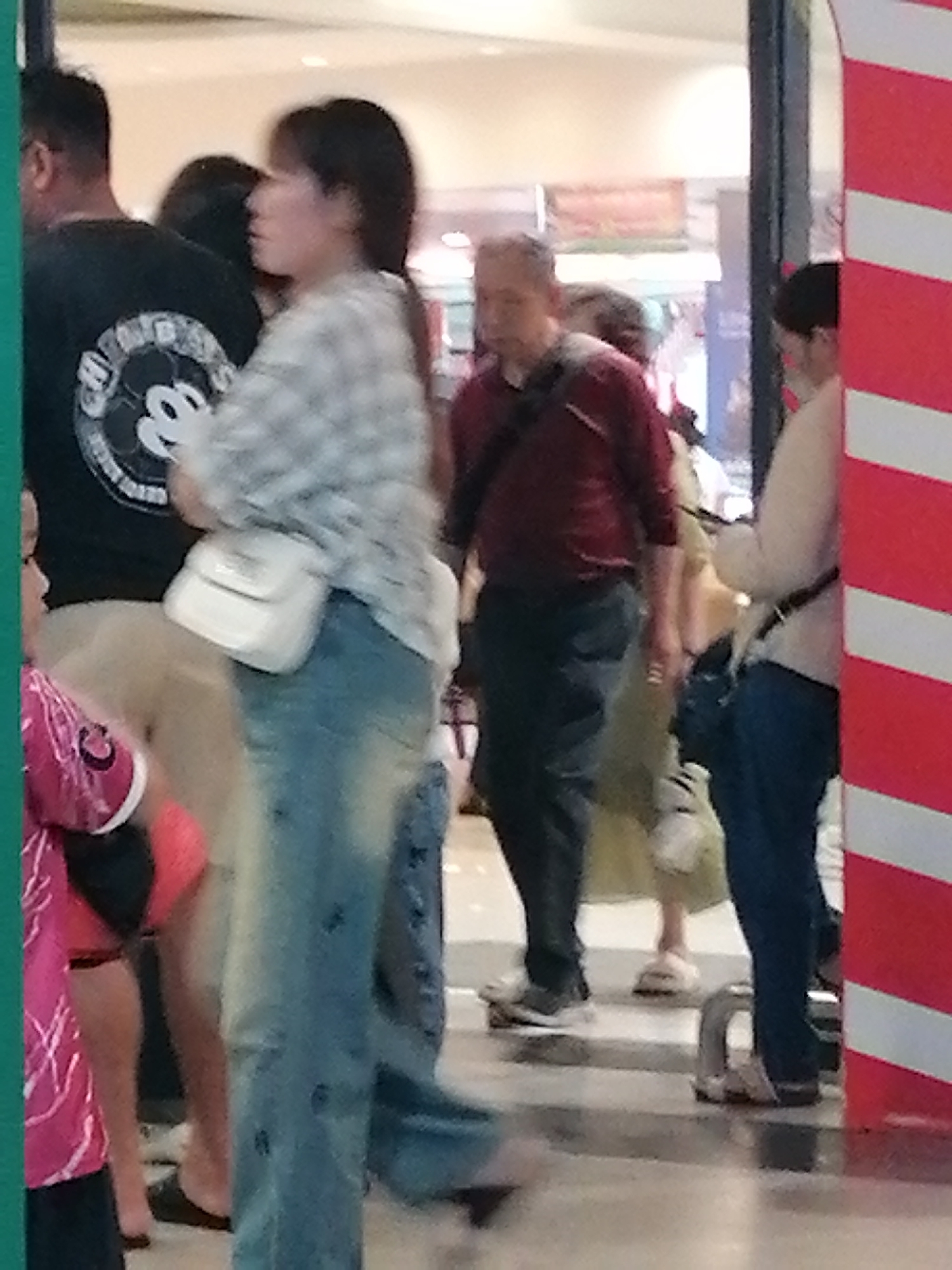 The image depicts a group of people in a public indoor space, likely a shopping mall or a similar commercial establishment, based on the presence of decorative elements like the red and white striped pillar and the ambient lighting. The scene is composed of several individuals, most of whom are partially obscured or out of focus, indicating a candid or casual photograph.
In the foreground, a woman is prominently featured, wearing a light-colored plaid shirt and blue denim jeans. She has a white crossbody bag slung over her shoulder. Her posture suggests she is walking or standing in a queue. To her left, a man wearing a black t-shirt with a circular graphic is visible, facing away from the camera. Further back, another man in a dark red long-sleeved shirt and dark pants is walking, with a bag strapped across his chest. A woman in a light-colored top and dark pants is partially visible to his right. In the background, other individuals and parts of the environment are blurred, suggesting movement and a relatively shallow depth of field.
The environment appears to be well-lit, with artificial lighting casting a soft glow. The background suggests retail displays or common areas within a shopping complex. There are no specific visual cues in the image that definitively place this scene in Da Lat, Vietnam. The architectural style, clothing, or any signage that would indicate a specific location in Da Lat are not discernible from the provided image.
